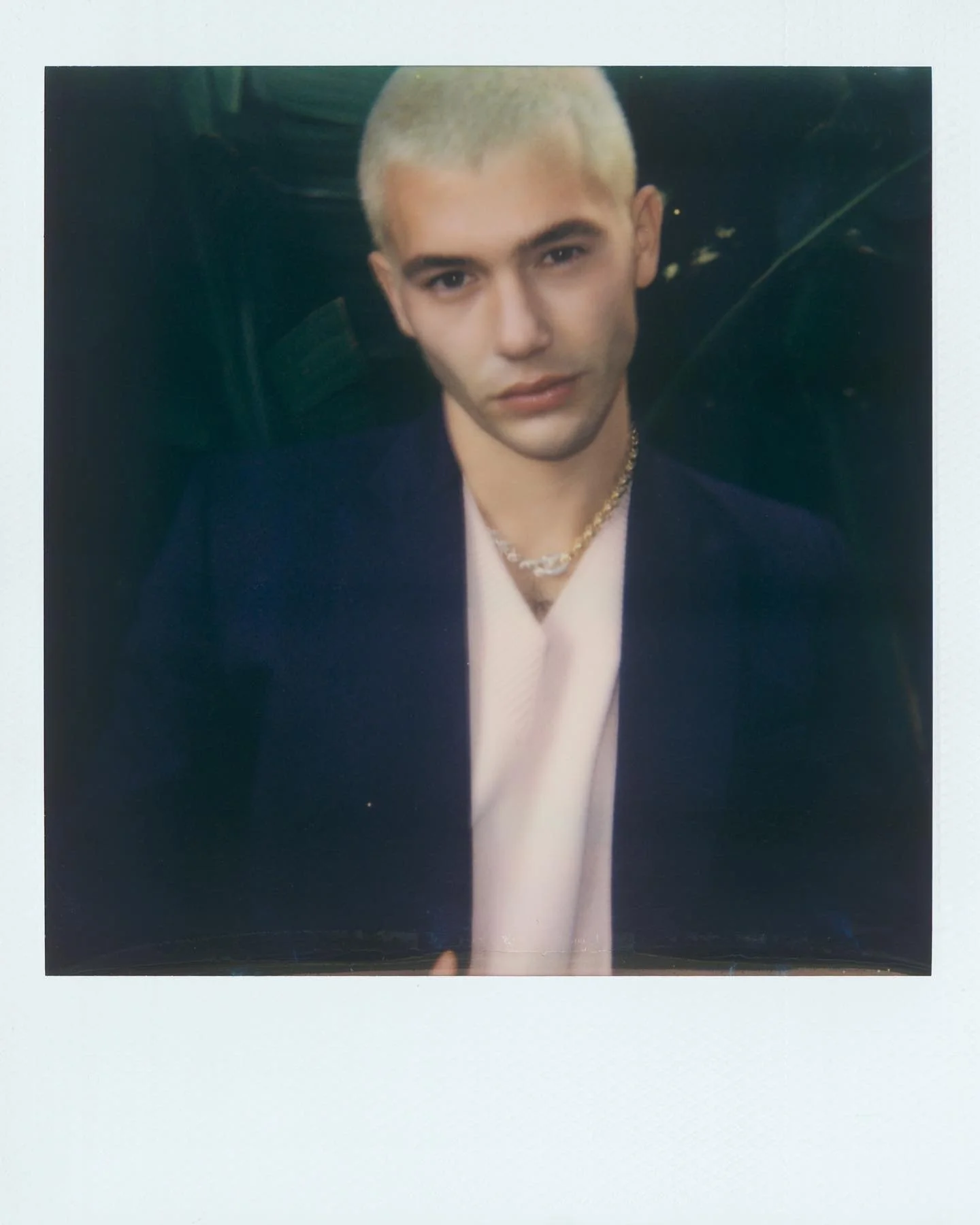 A young person with platinum blond hair wearing a dark blazer, light pink shirt, gold chains, and looking at the camera with a serious expression.