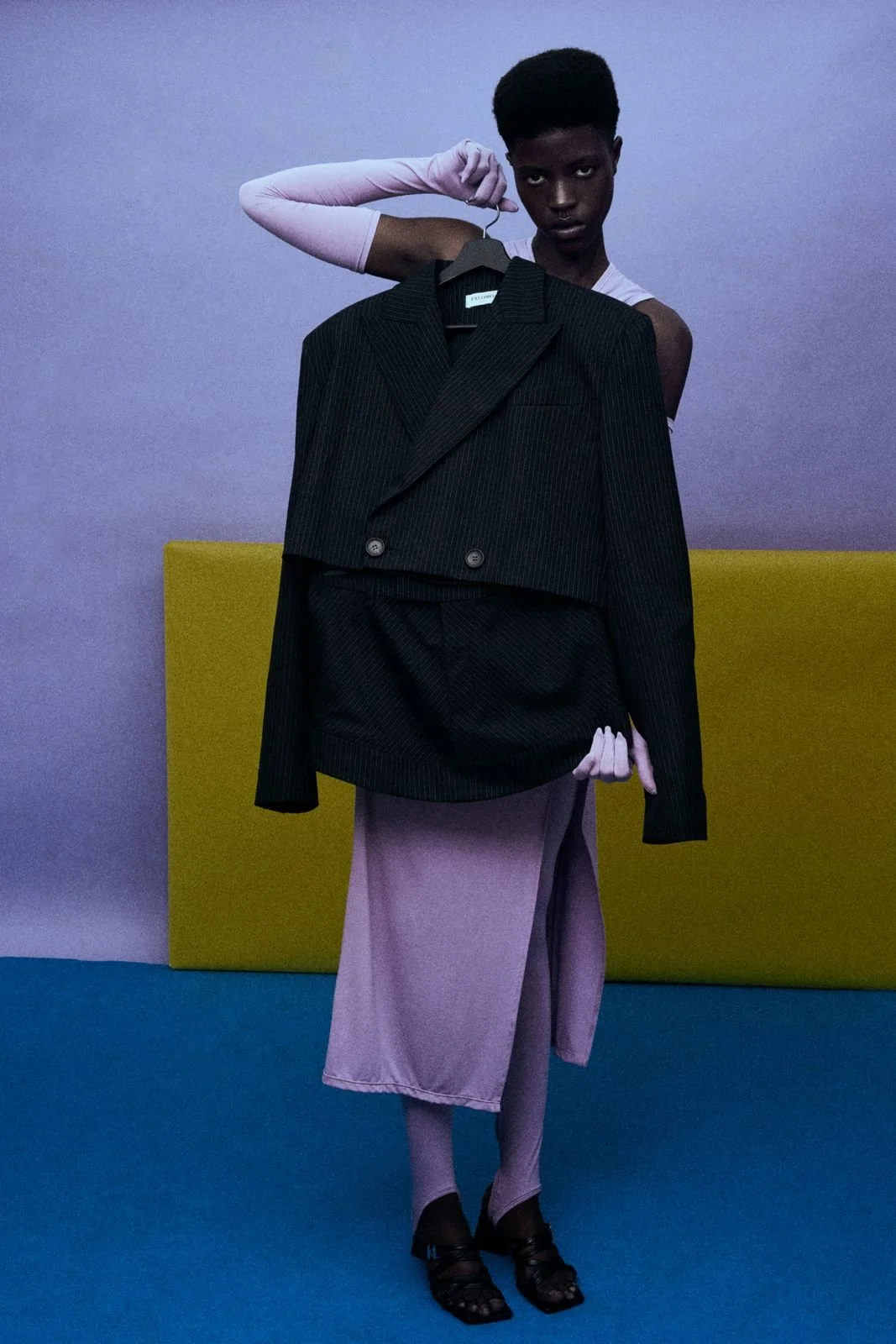 A person with dark skin and short hair stands in front of a multicolored background, holding a black pinstripe blazer on a hanger. The person wears a sleeveless top, a long lavender skirt, and black heels, with one pink-gloved hand holding the blazer