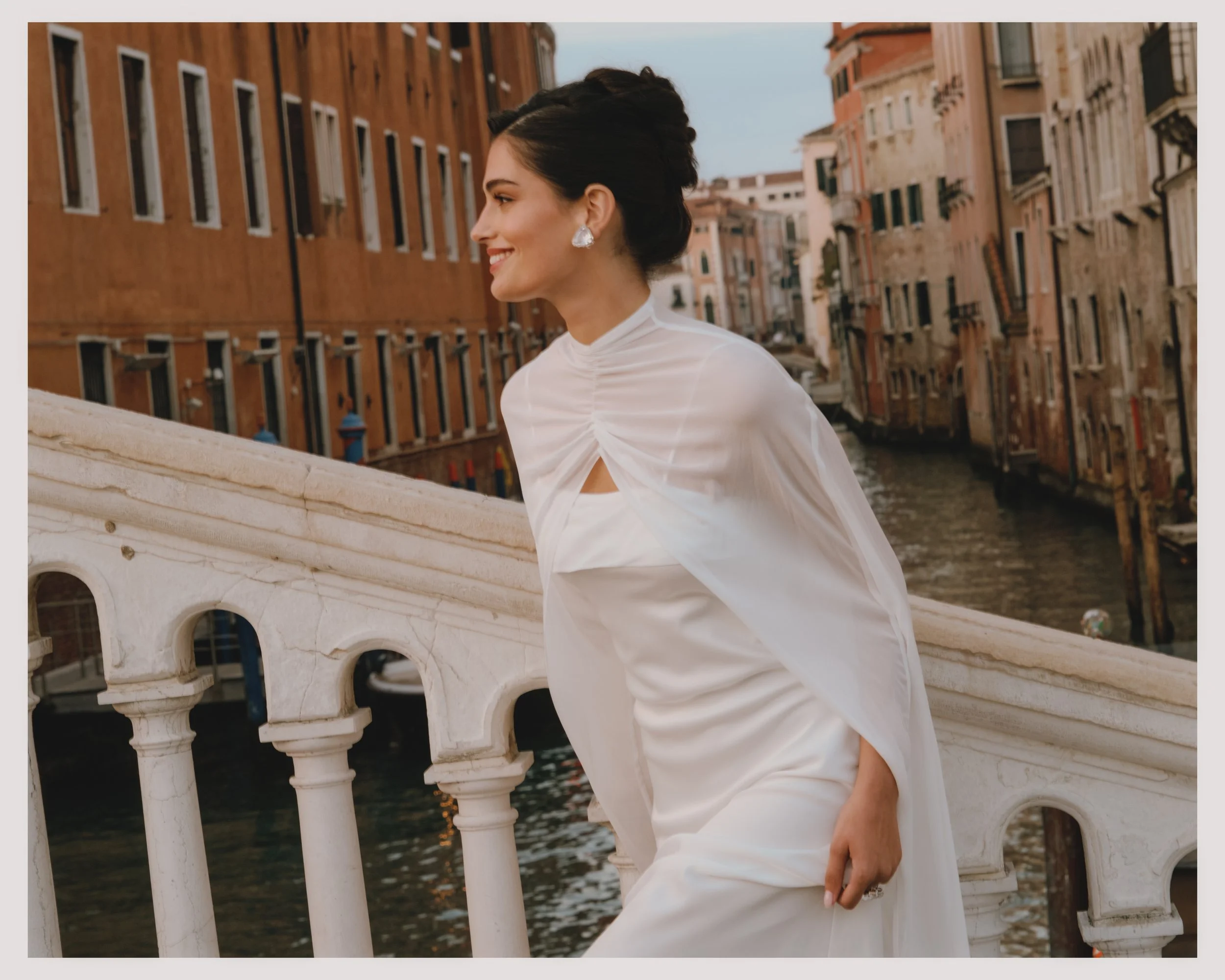 A woman in a white dress and earrings standing on a bridge over a canal in Venice, Italy, with historic buildings on either side.
