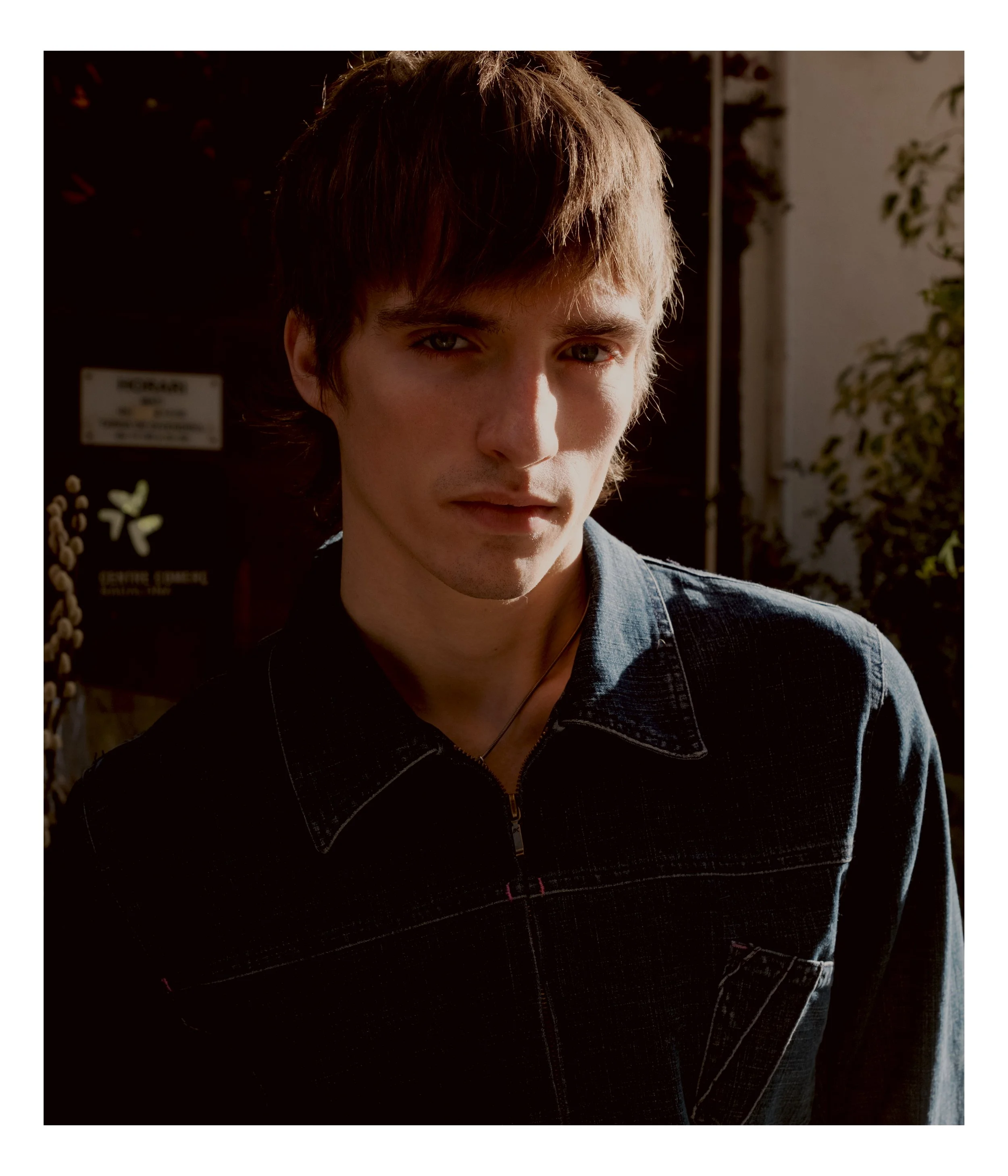 Young man with brown hair and a serious expression, wearing a dark denim jacket outdoors during daytime.