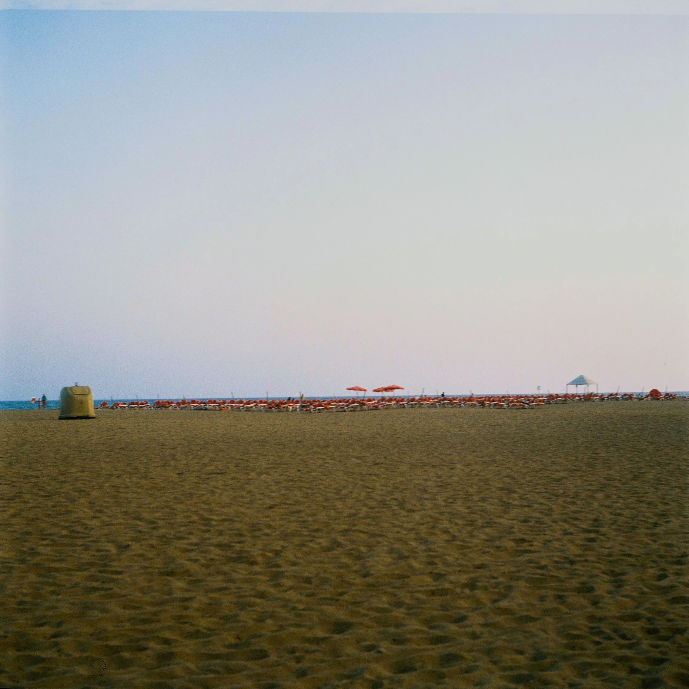Empty sandy beach with lounge chairs and umbrellas near the water, with a small pavilion structure in the distance under a pale sky.