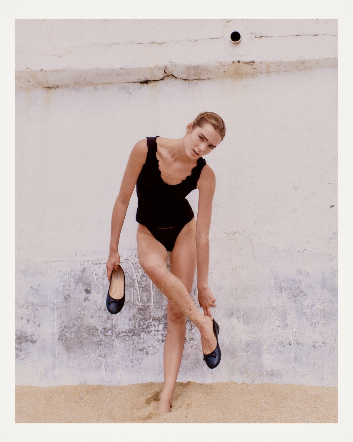 A woman in a black top and underwear standing on sand against a white wall, holding her shoe and adjusting her ankle.