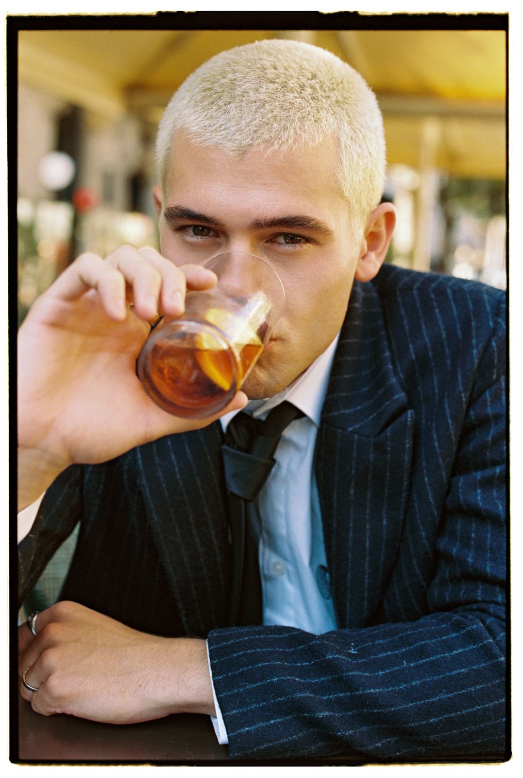 Man with bleached blond hair wearing a navy pinstripe suit and white shirt, drinking a glass of whiskey at an outdoor cafe.