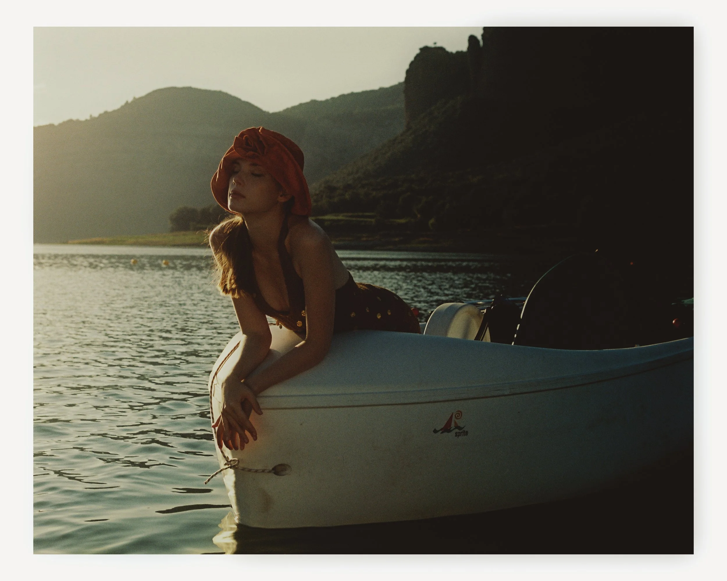 A woman with long hair wearing a red headwrap and dark dress, leaning on a small rowboat on a body of water during sunset with mountains in the background.