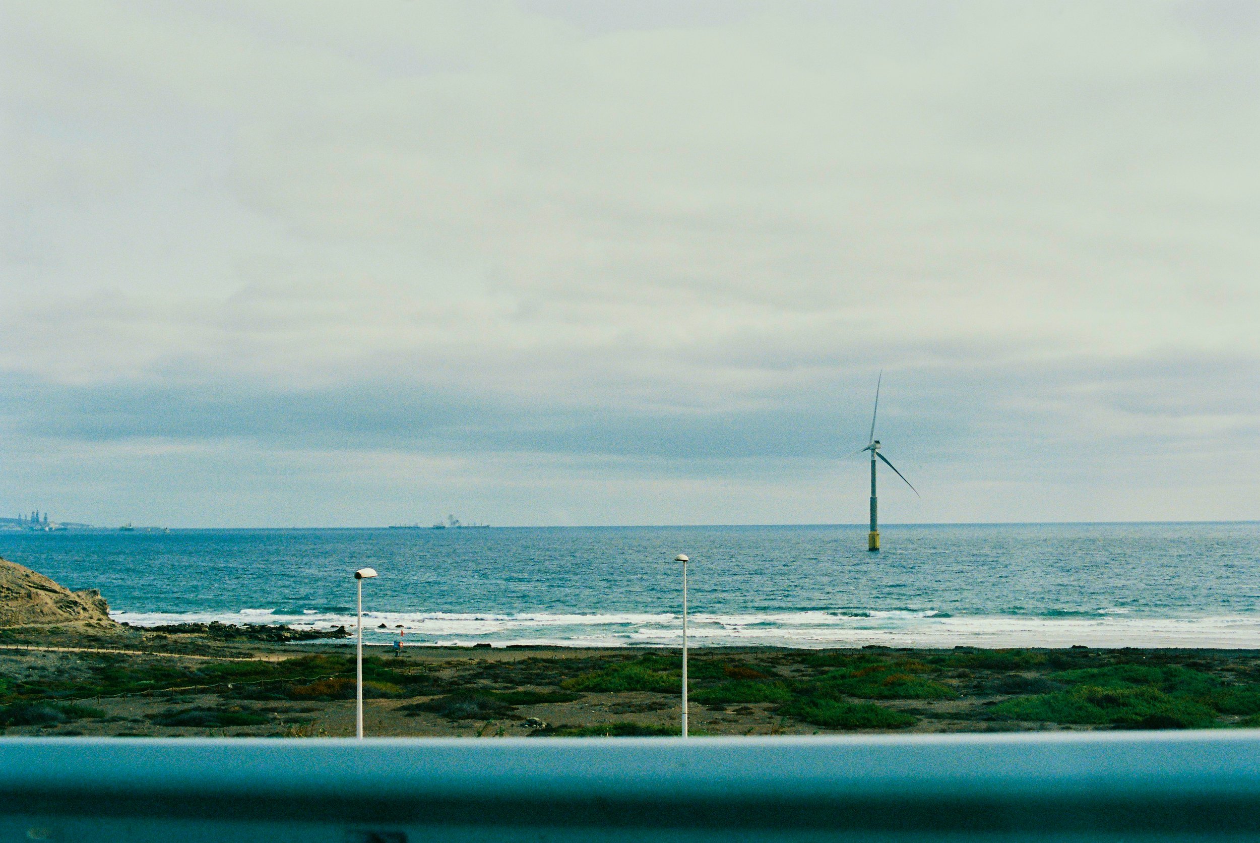 A coastal scene with a wind turbine in the ocean, two streetlights on the shore, rocky terrain, and cloudy sky.