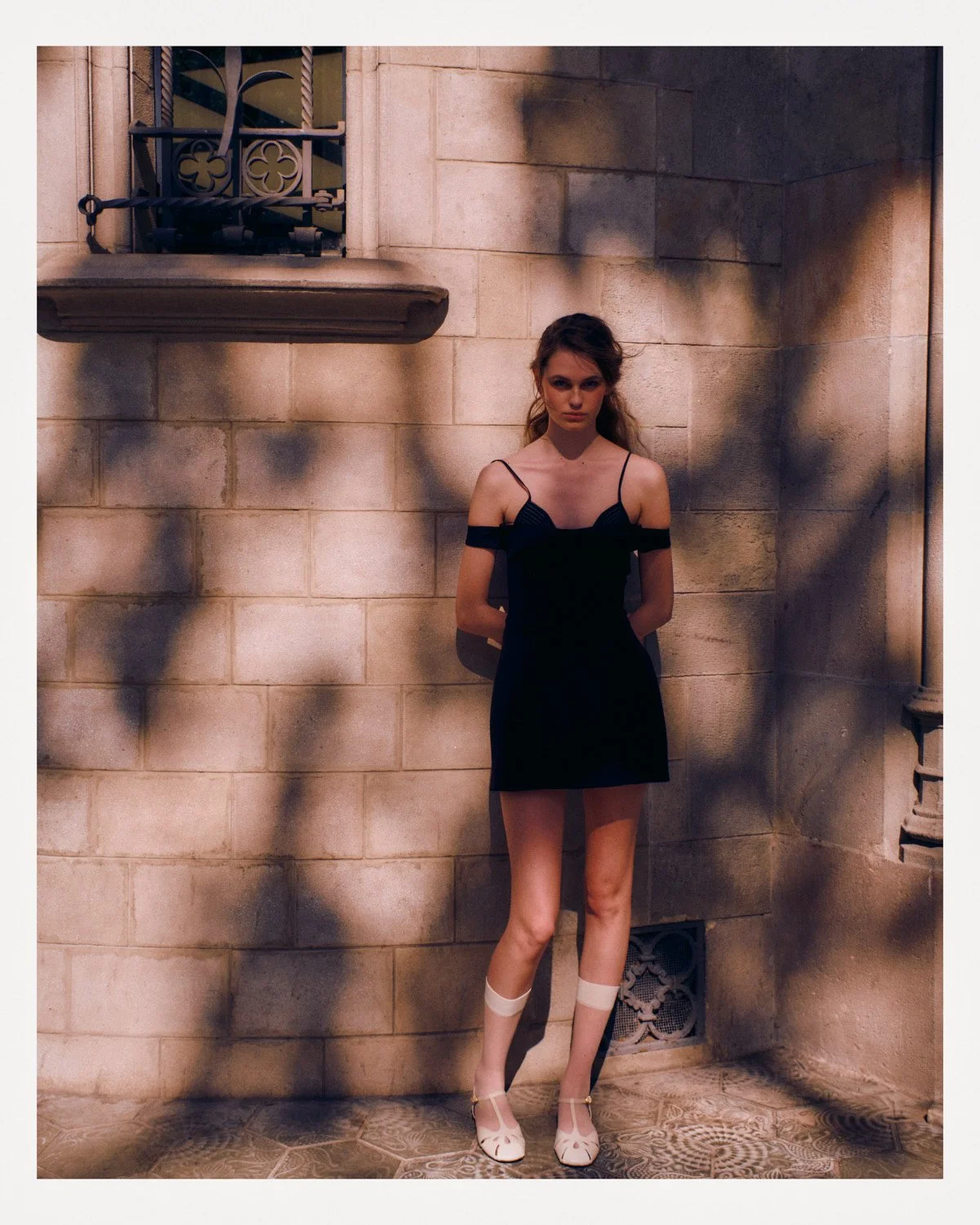 A young woman in a black dress with spaghetti straps standing against a beige stone wall with shadows of leaves, beside a window with decorative wrought iron bars.