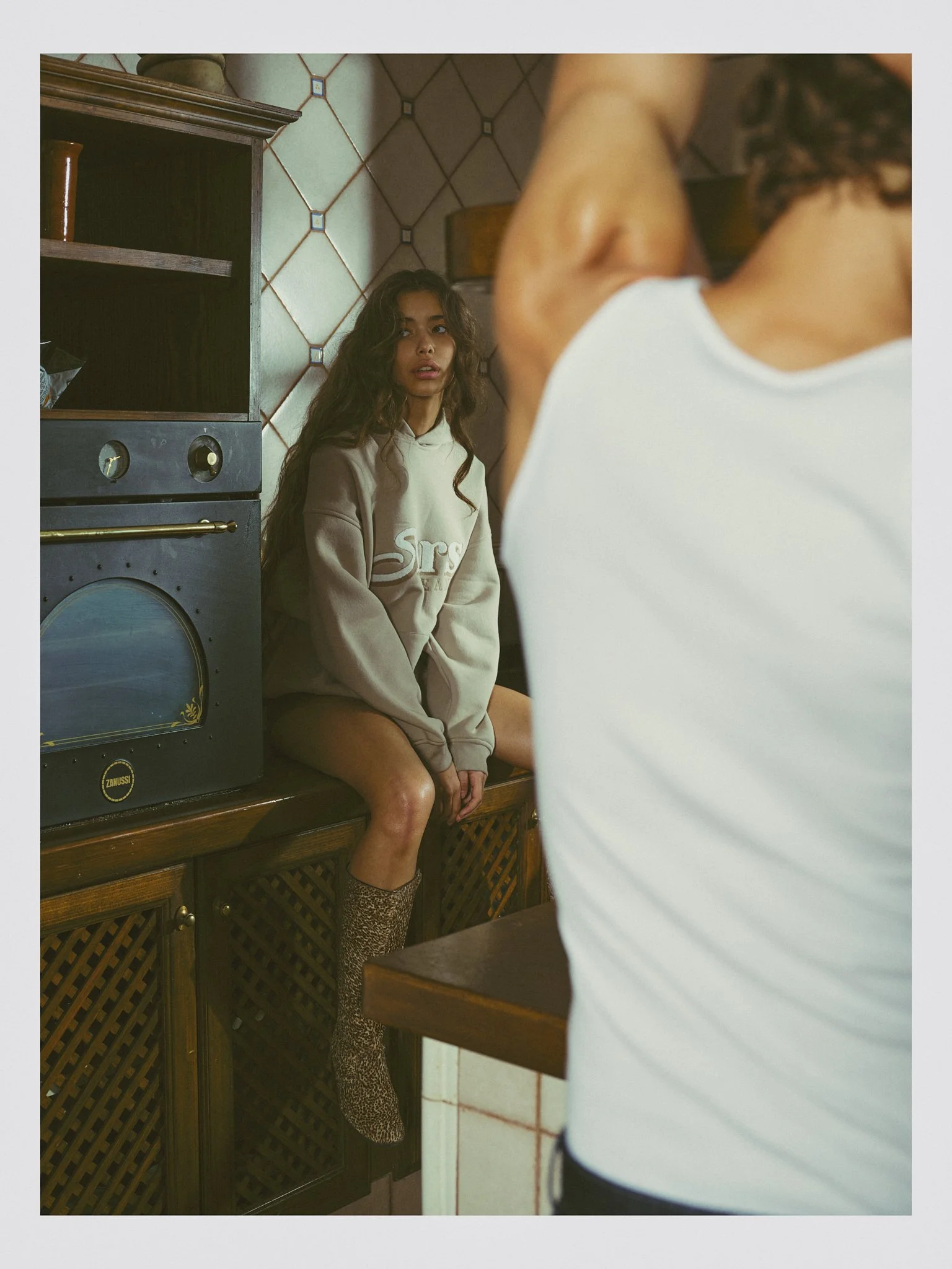 A woman sitting on a wooden cabinet in a kitchen, looking at a person in a white tank top in front of her. The woman wears a beige hoodie and leopard print socks, with long wavy hair, and the kitchen has tiled walls and an oven.