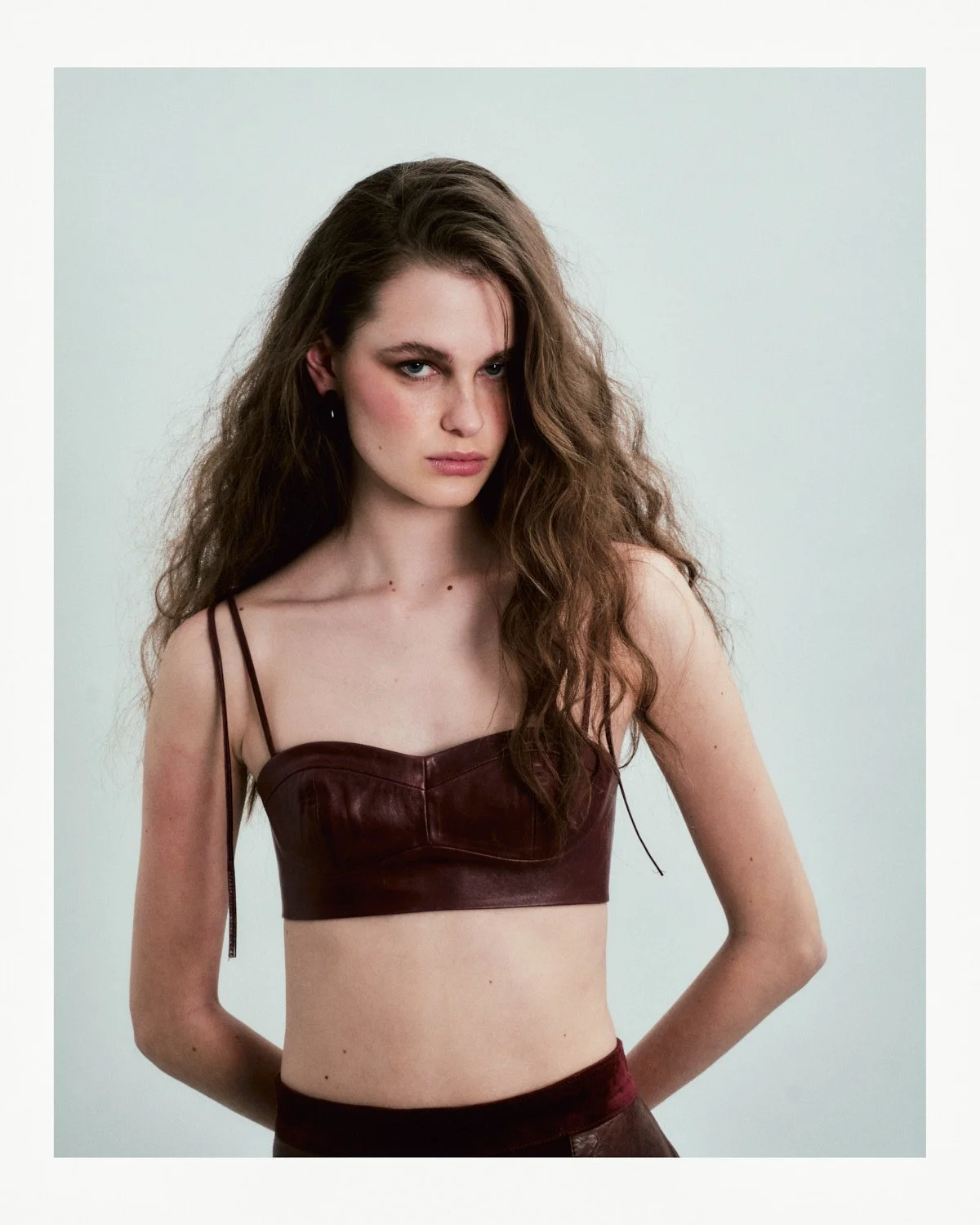 A young woman with long, curly brown hair and blue eyes poses against a plain light background. She wears a dark red leather crop top with spaghetti straps and matching high-waisted bottoms.