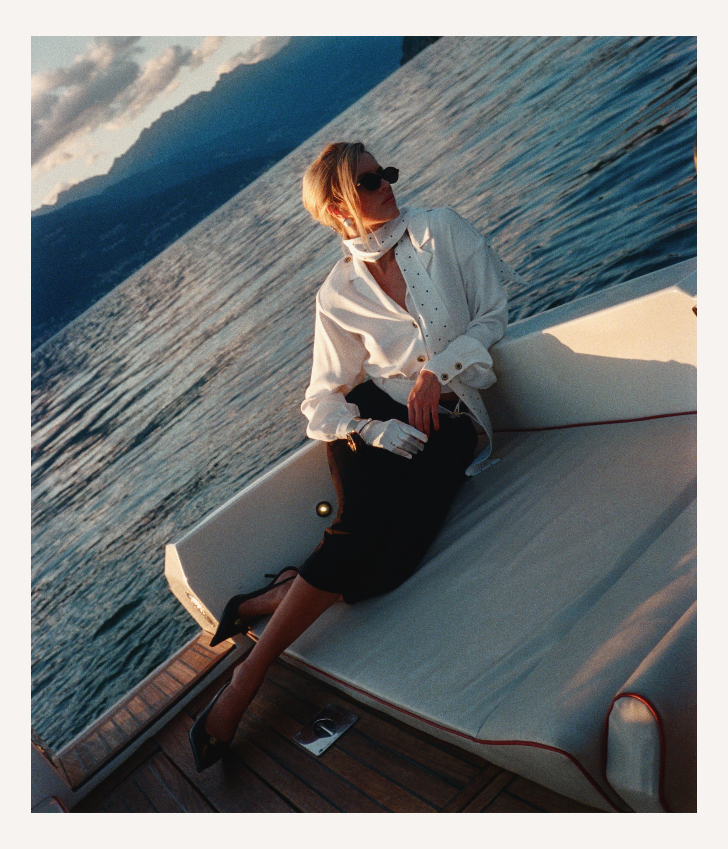 A woman with blonde hair wearing sunglasses, a white blazer, black skirt, and black heels sitting on the deck of a boat at sunset, with mountains and water in the background.