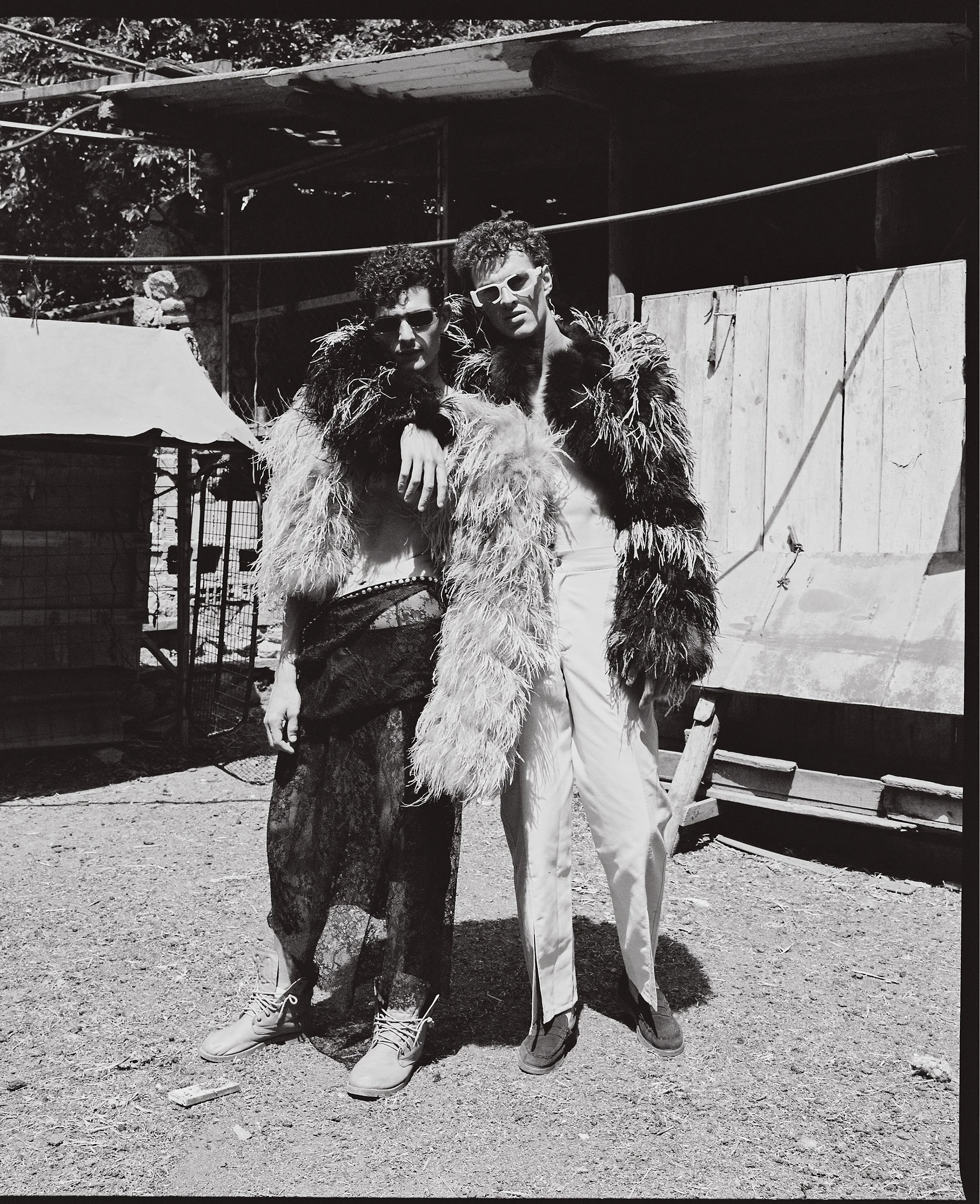 Two people standing outdoors, both wearing large, furry coats and sunglasses. One person is wearing a floral skirt and boots, while the other is dressed in light-colored pants and shoes. The background includes a makeshift wooden structure and a wire