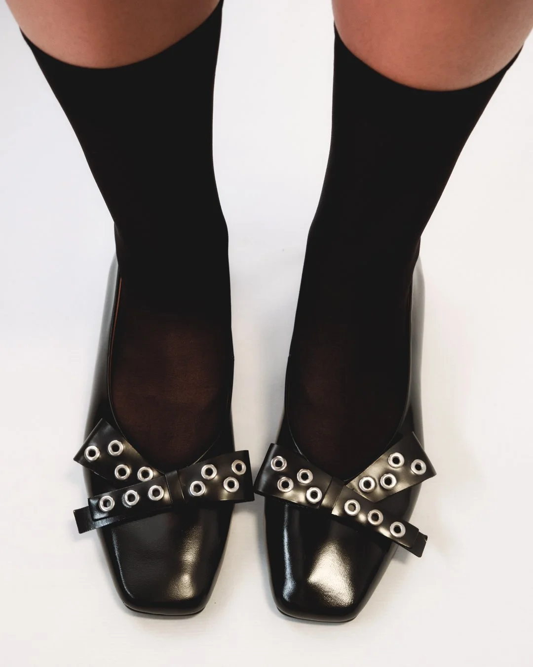 Pair of black shoes with bows and metal grommets, worn with black stockings.