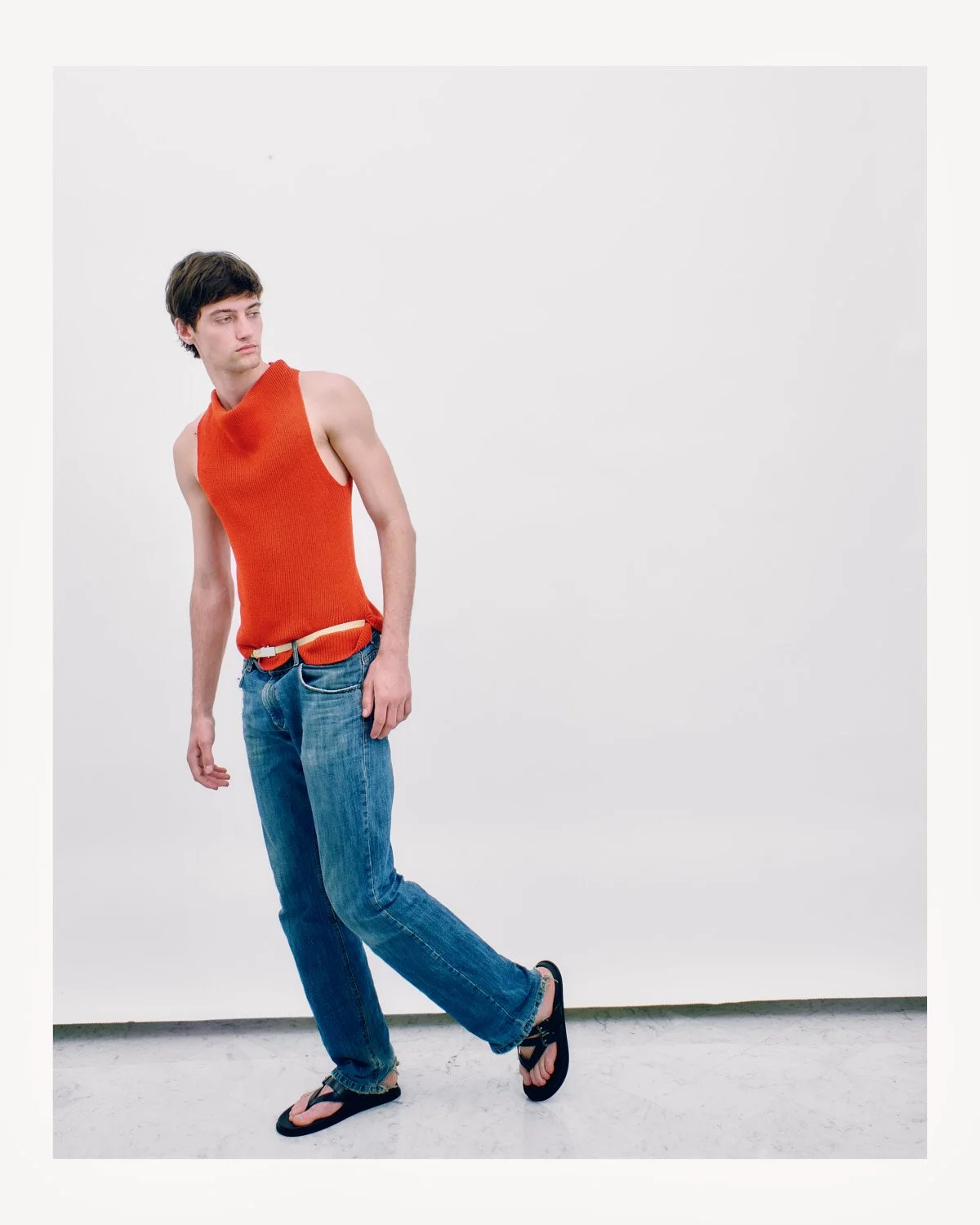 Young man with dark hair wearing an orange sleeveless knit top, blue jeans, and black sandals standing against a plain white background.