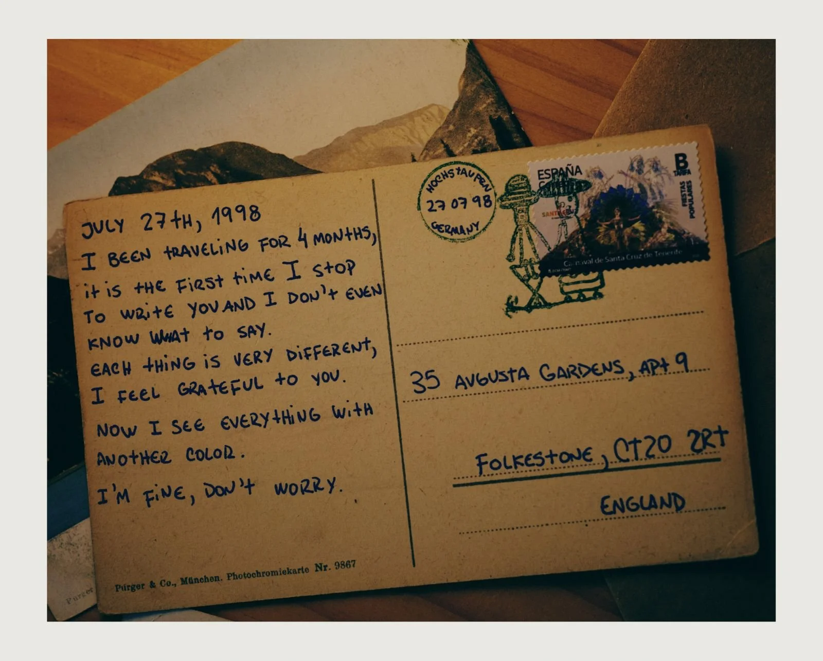 A vintage postcard with handwritten note, a stamp from Spain, and a colorful illustrated stamp featuring a landscape and figure. The note is dated July 27, 1998, and describes a four-month travel experience.