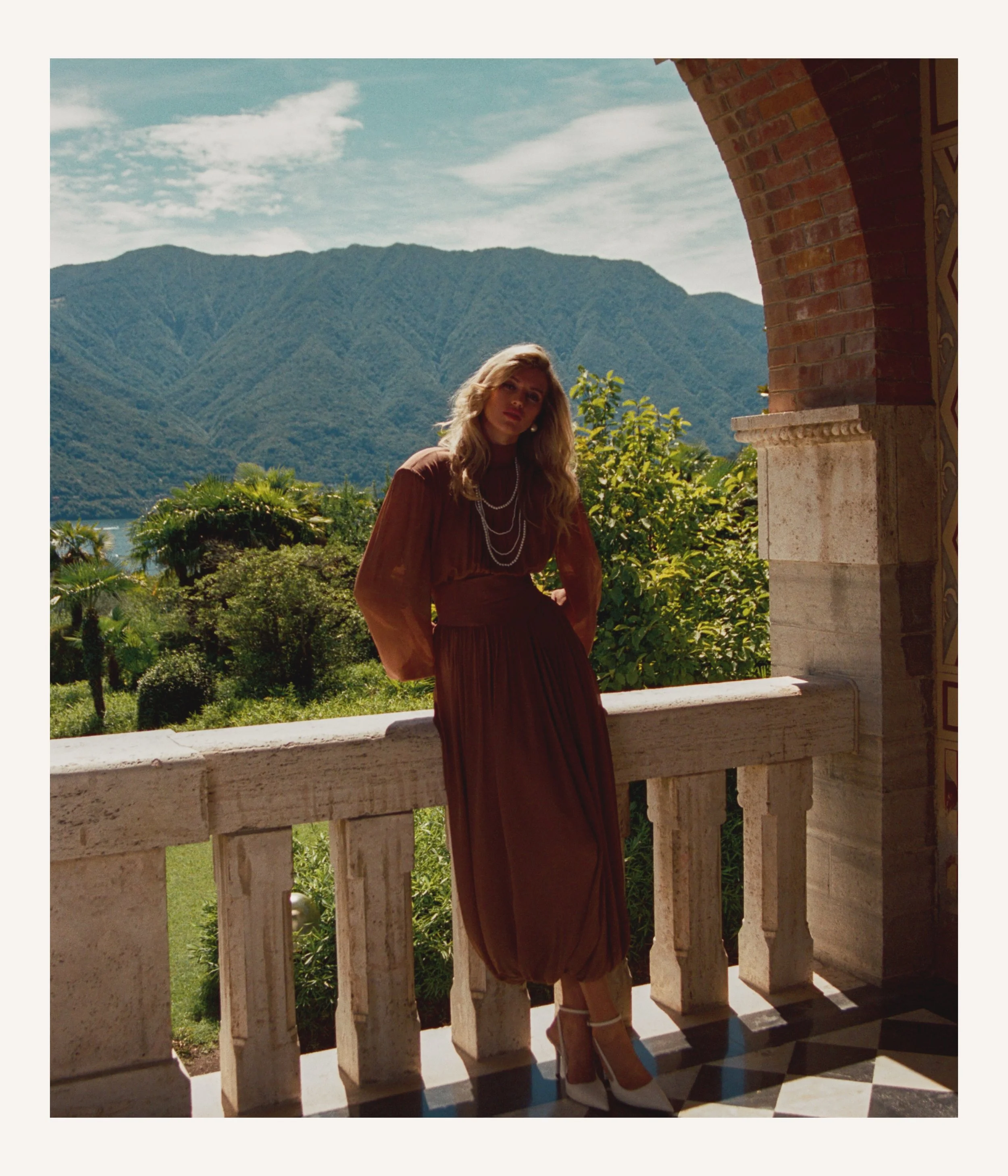 A woman in a brown dress and high heels standing on a balcony with a mountain landscape in the background.