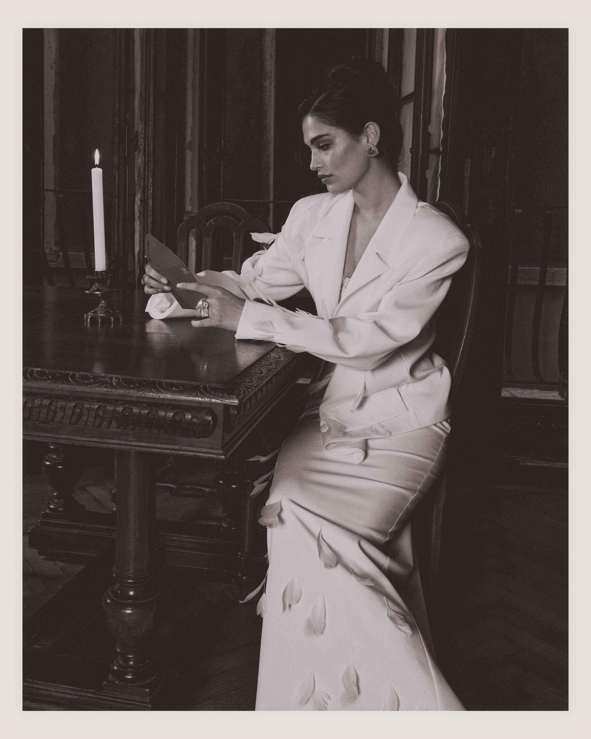 A woman in a white suit and dress sitting at a wooden table with a lit candle, looking at a photo or card she is holding.