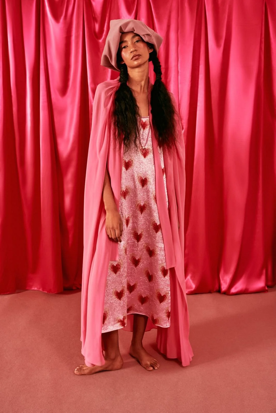 A woman standing barefoot in front of a pink satin curtain, wearing a pink dress adorned with red hearts, a long pink coat, and a pink head wrap.