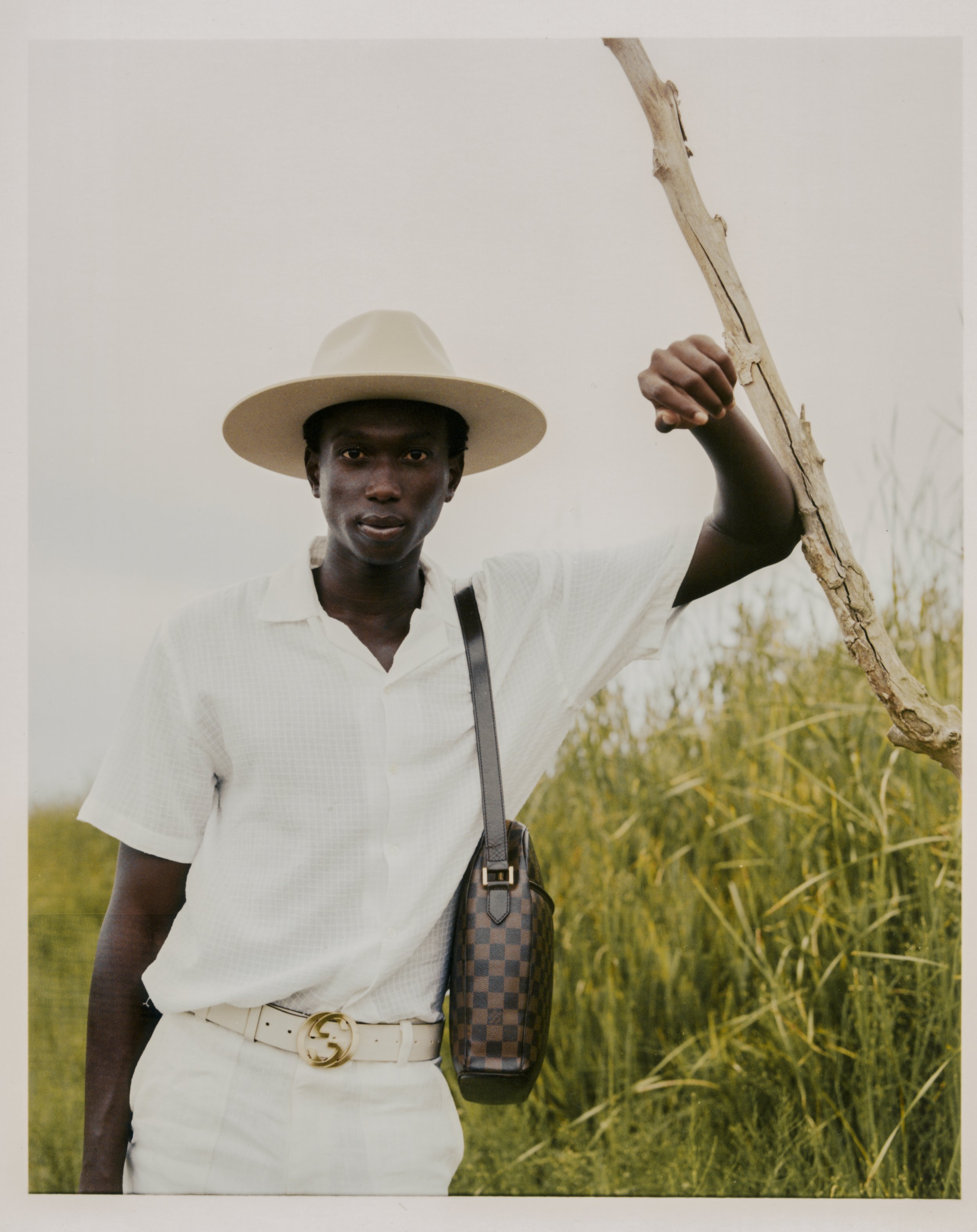 A young man in light-colored clothing and a wide-brimmed hat standing outdoors in a field, holding a large stick.