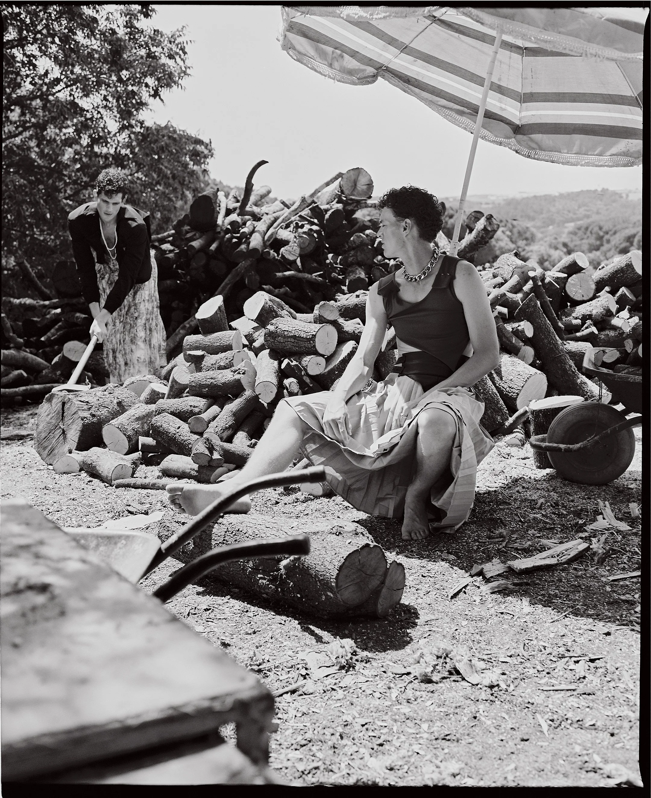 A black-and-white photo of a woman sitting on a log under a large striped umbrella, with a large pile of chopped firewood behind her. A person in the background is chopping wood with an axe.