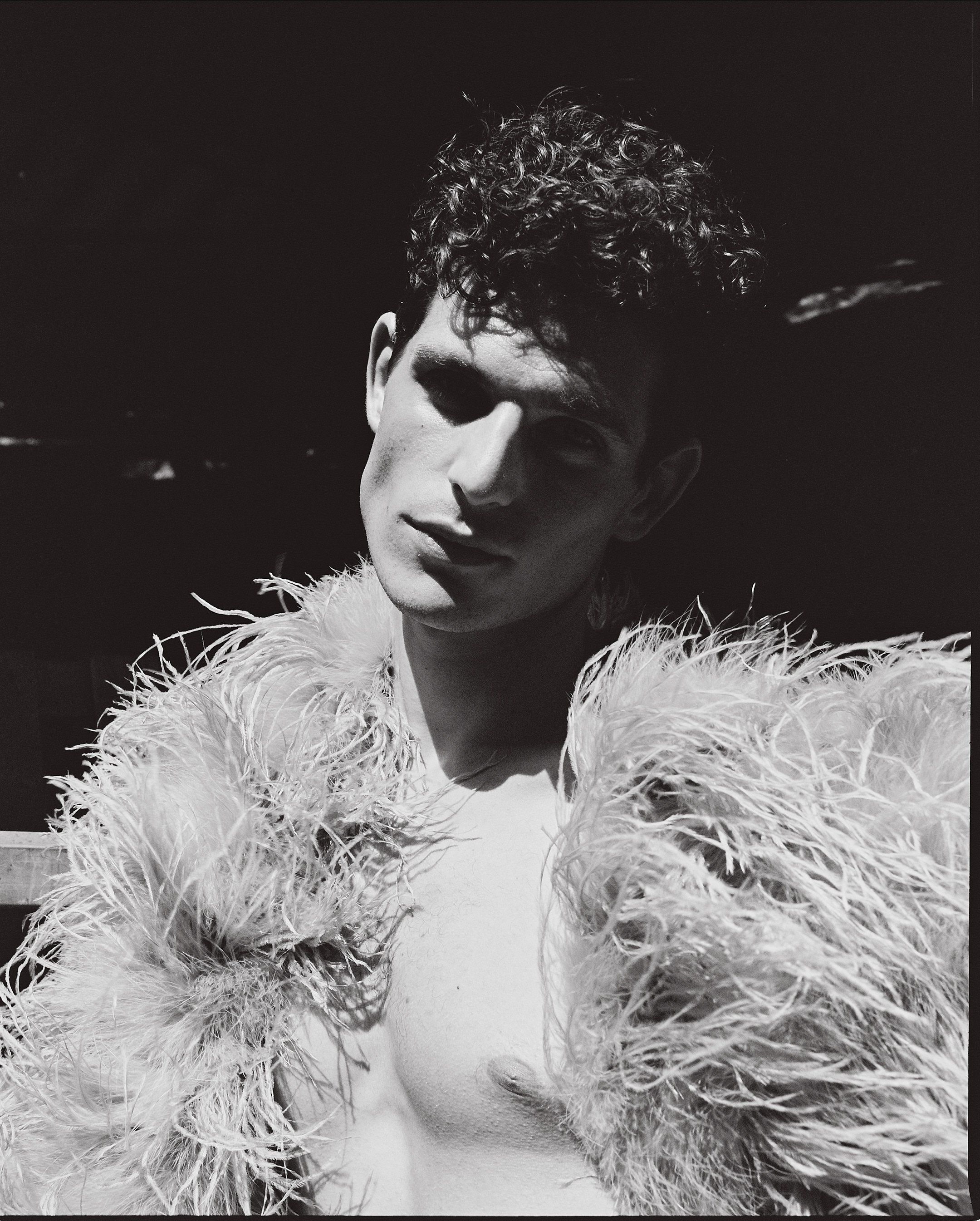 Black and white photo of a young man with curly hair wearing a feathered jacket, looking at the camera with a confident expression.