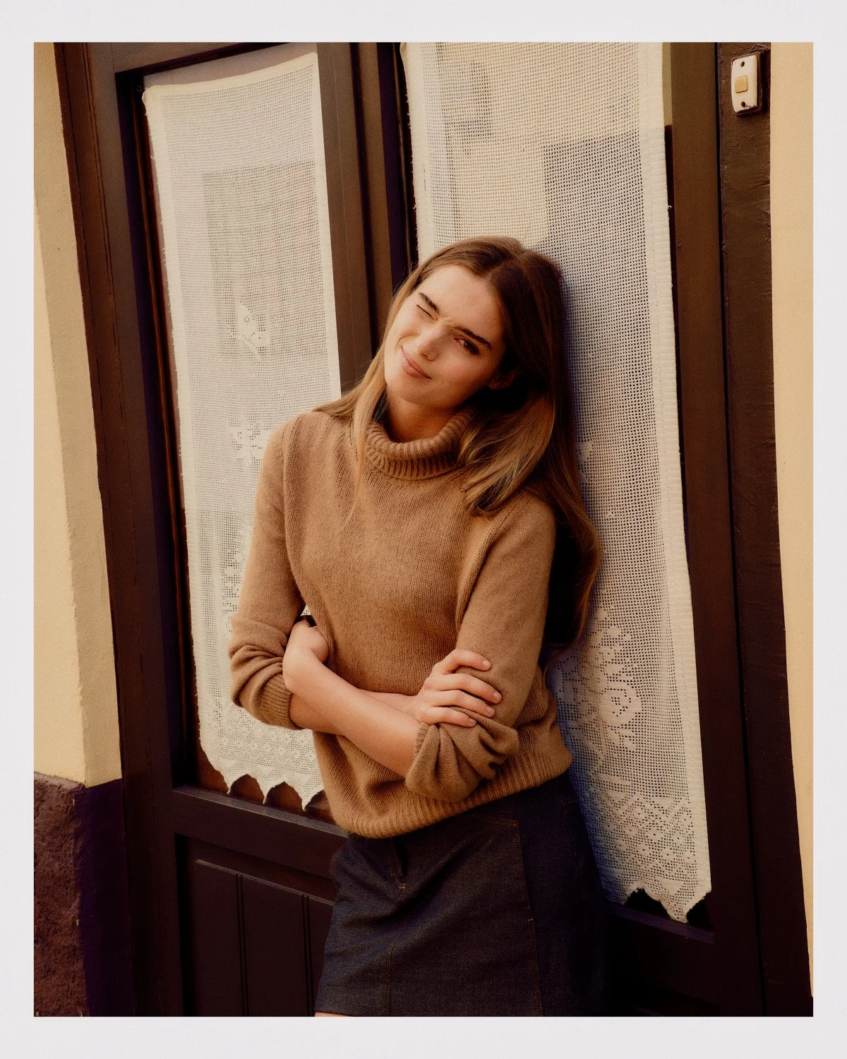 Young woman with light brown hair wearing a tan sweater, standing in front of a window with lace curtains, smiling and winking.