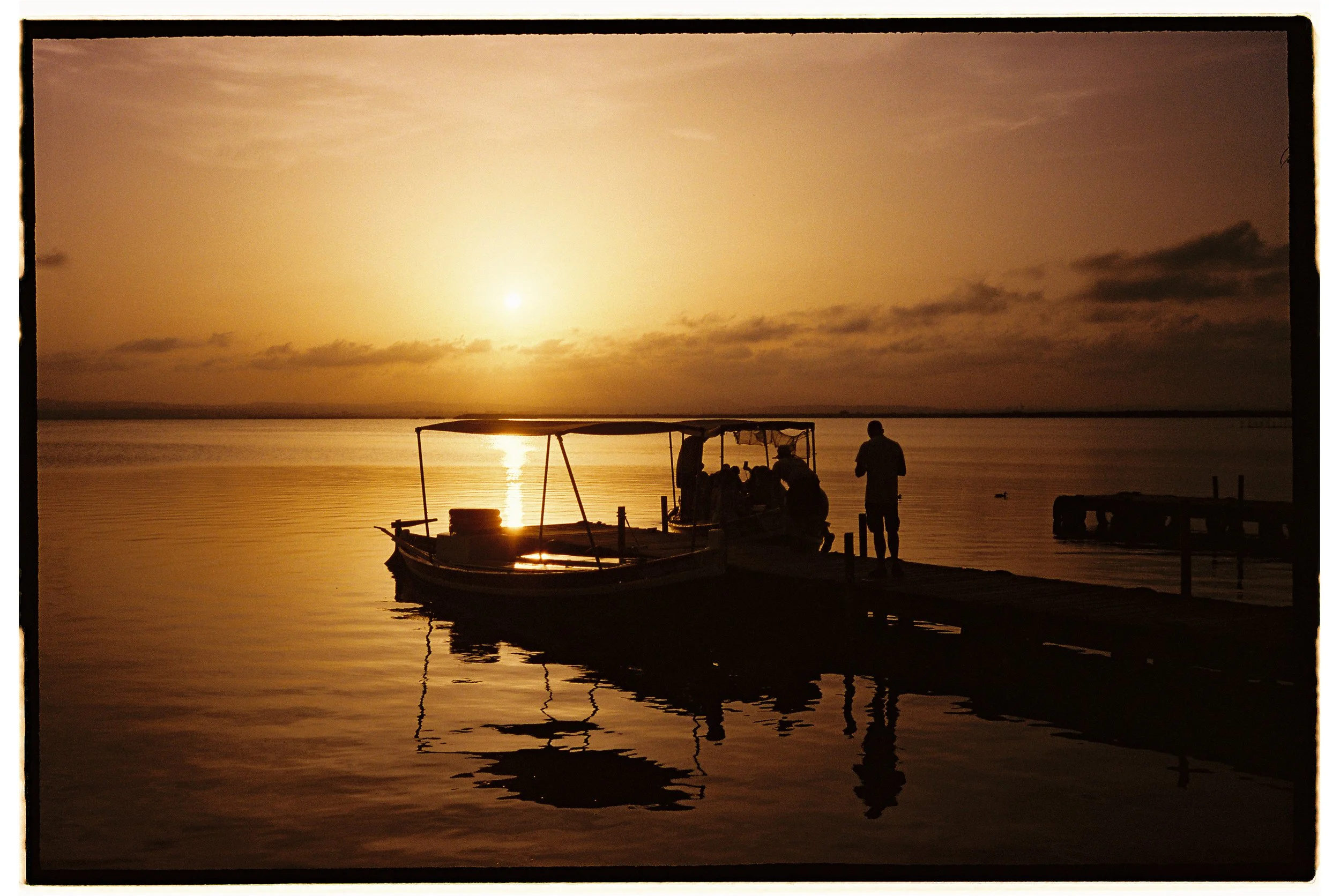 Silhouette of a boat docked at a pier with people around it during sunset or sunrise over calm water with a partly cloudy sky