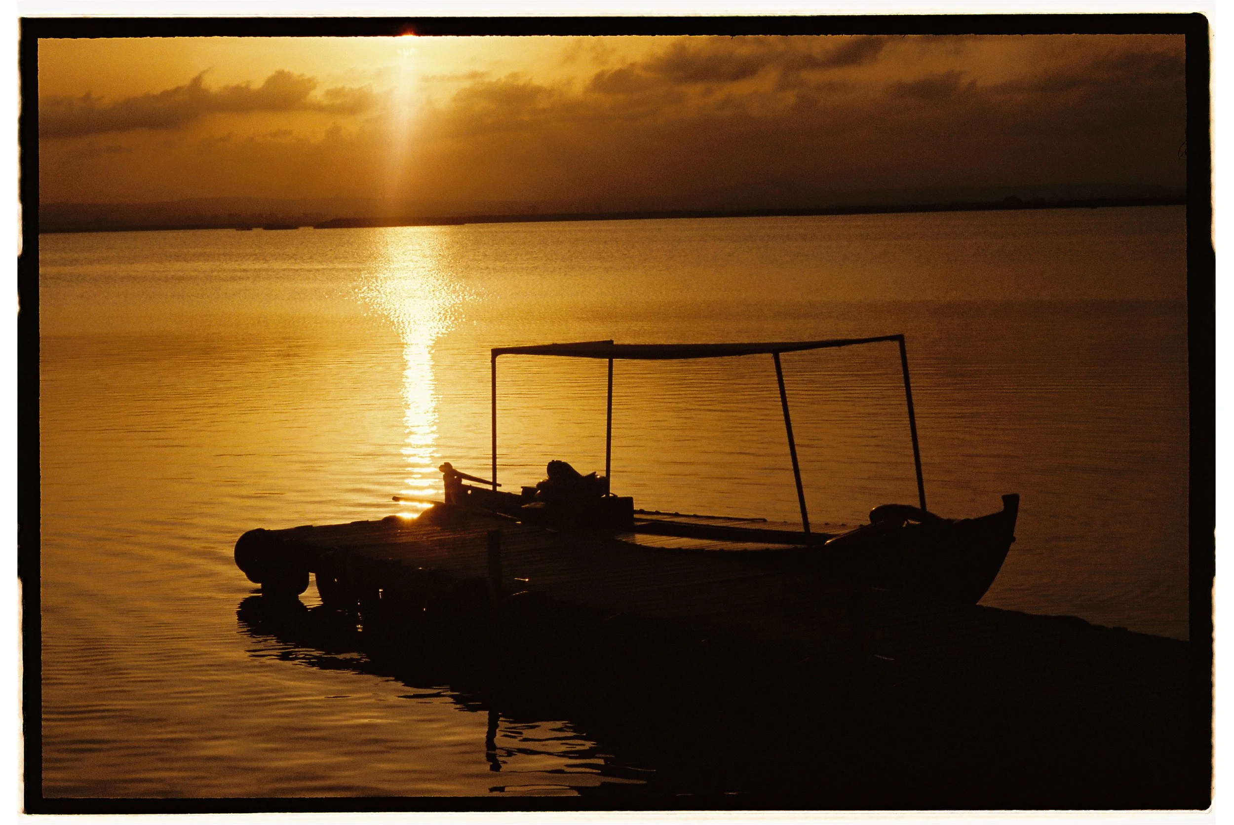 Silhouette of a small wooden boat on calm water during sunset or sunrise, with a reflection of the sky and clouds on the water.