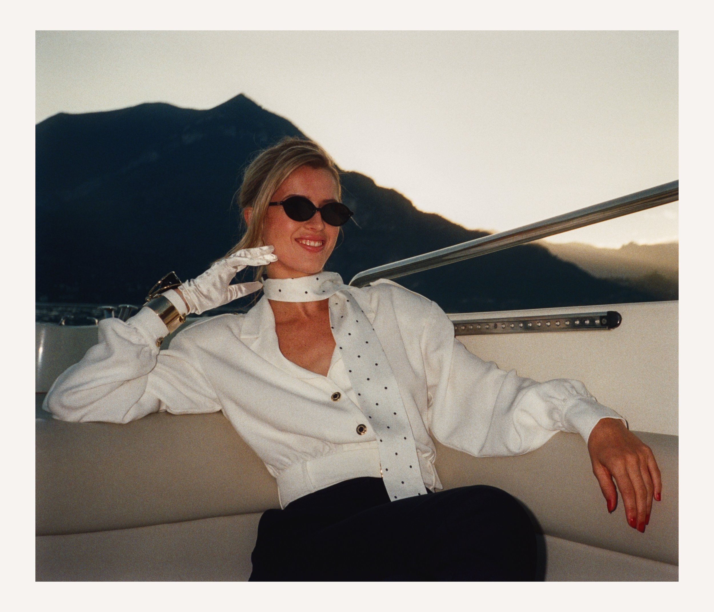 Woman with sunglasses, white blouse, and polka dot scarf relaxing on a boat with mountains in the background at sunset.