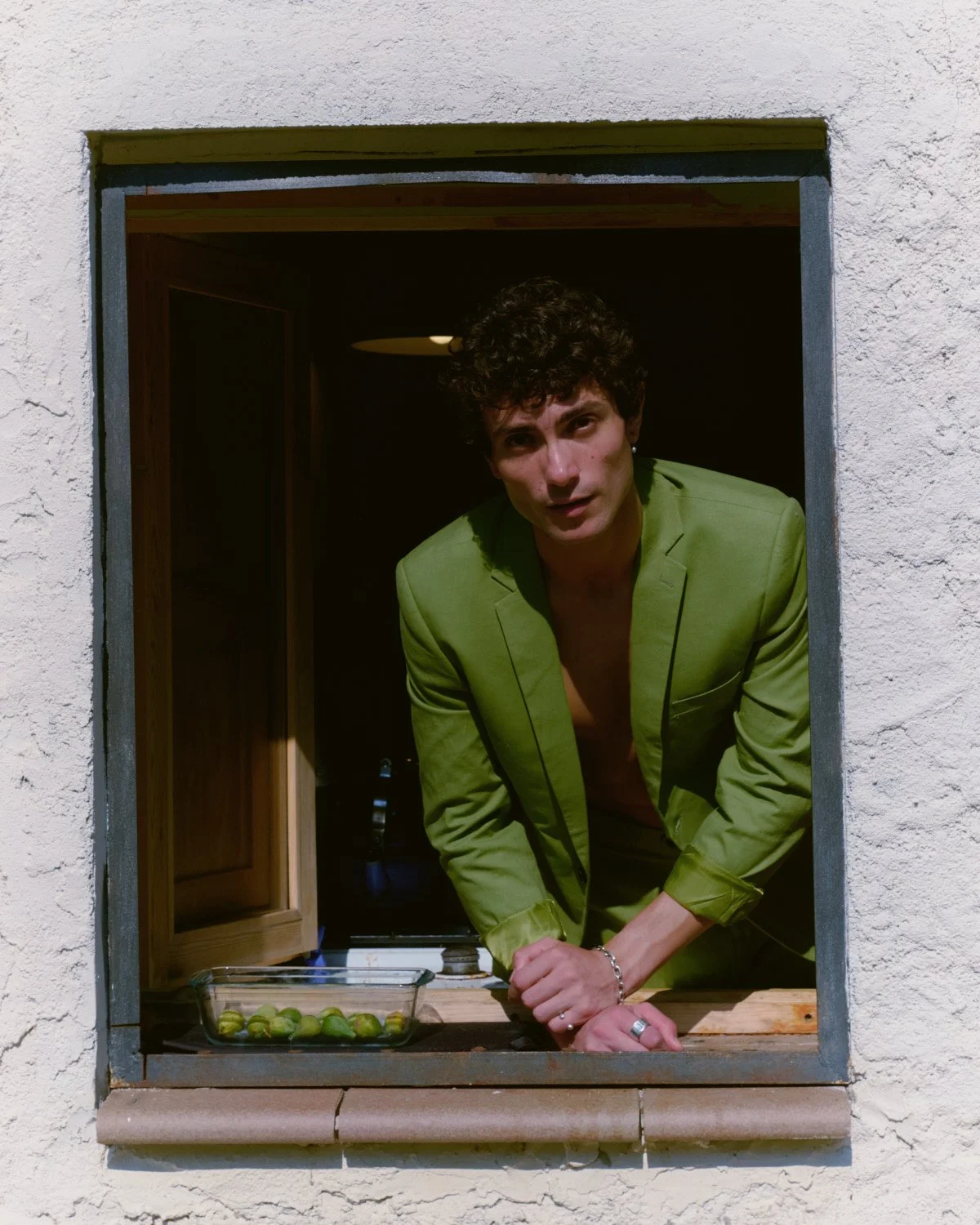 A person with dark curly hair wearing a green blazer leaning out of a window with a white stucco wall, with a small glass dish of fresh figs on the windowsill.