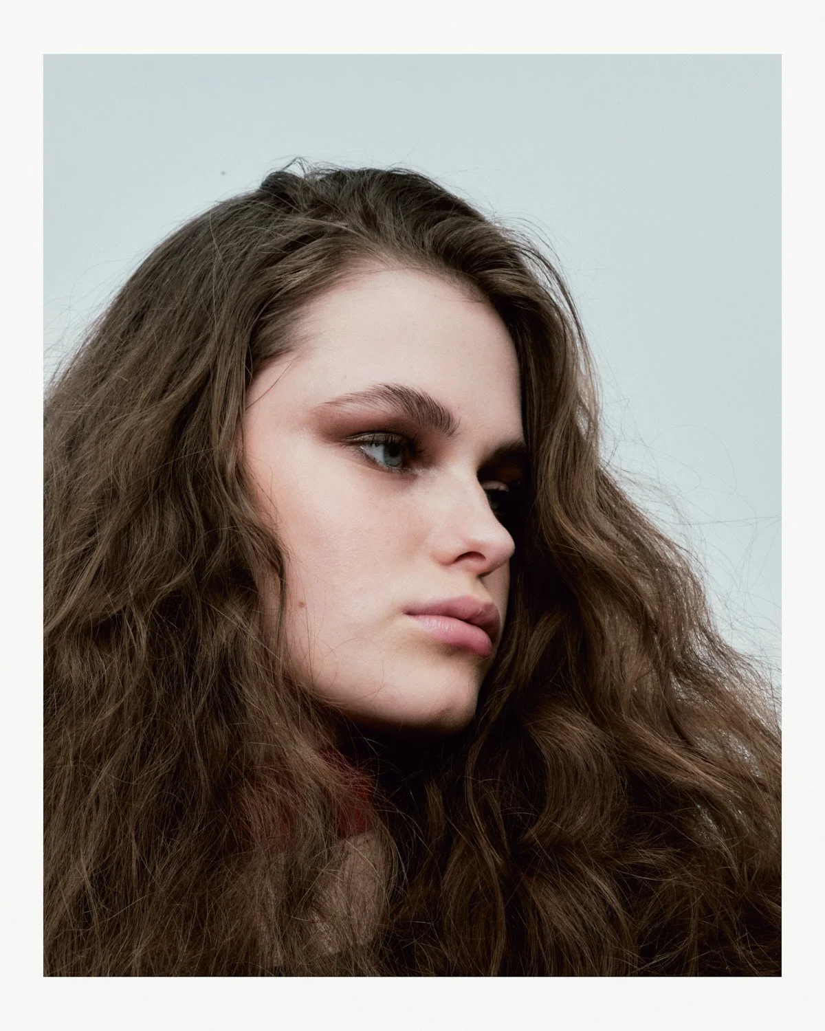 Close-up portrait of a young woman with long, curly brown hair, fair skin, and wearing makeup with brown eyeshadow.