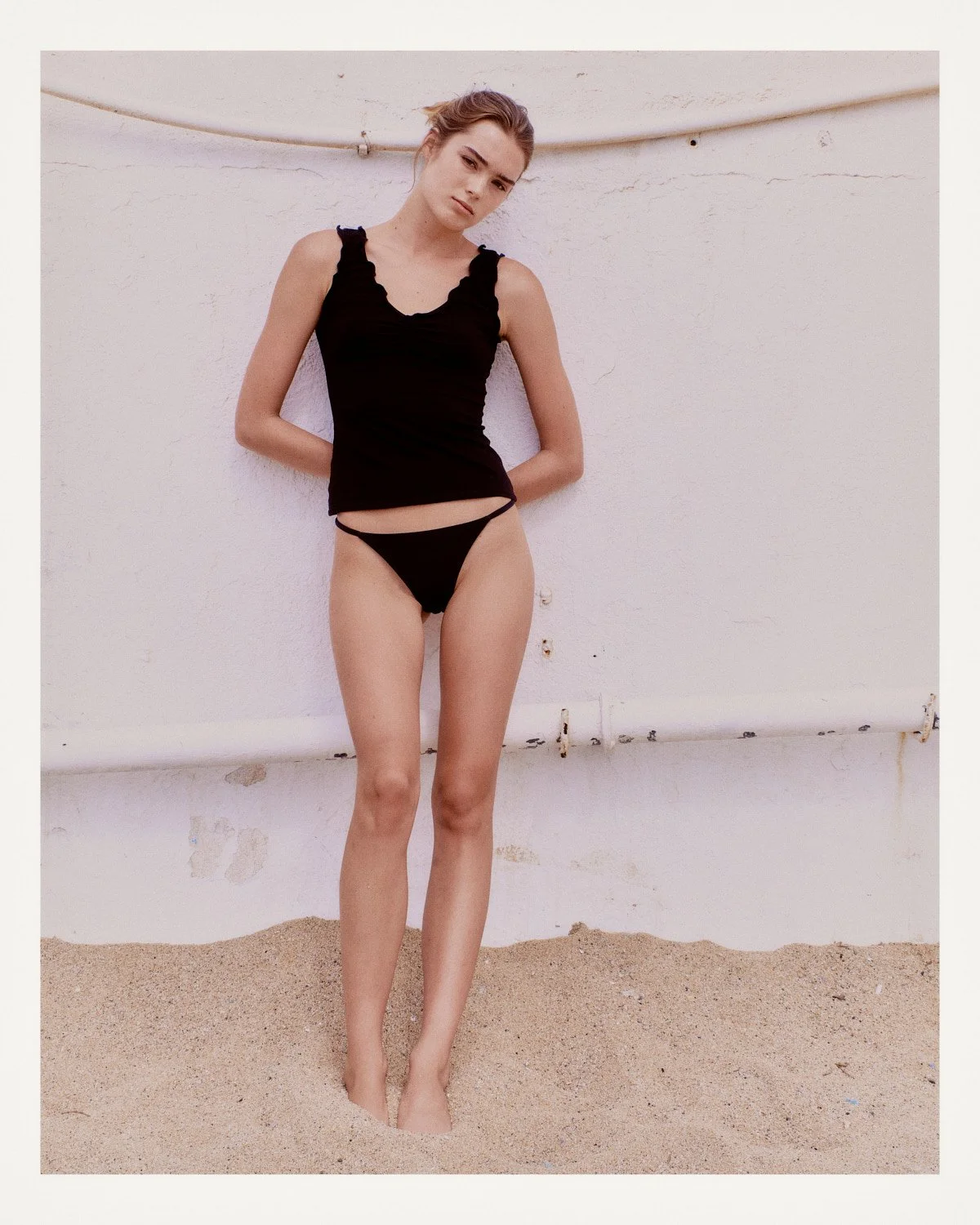 Young woman in black tank top and underwear standing barefoot on sandy ground, leaning against a white wall outdoors.