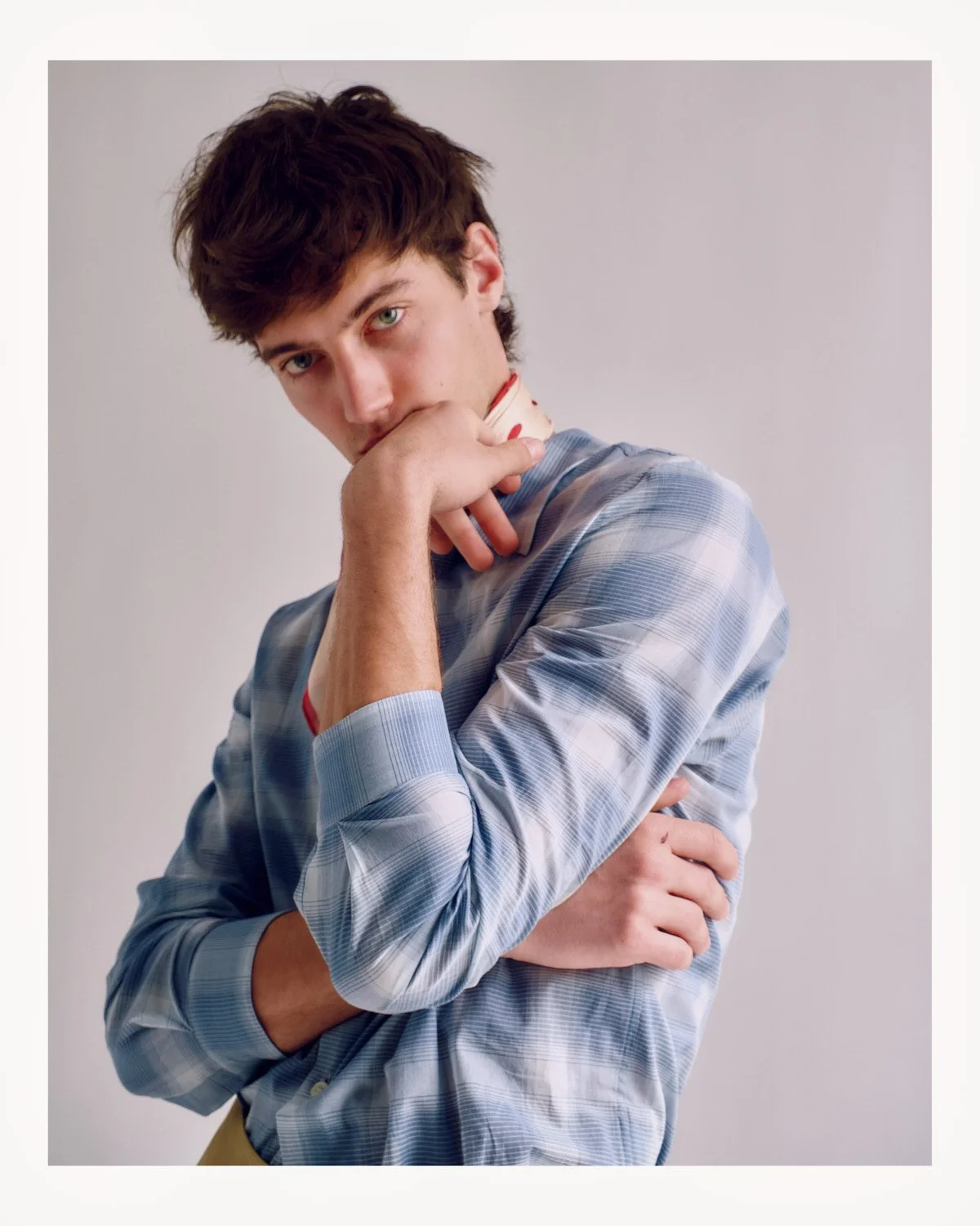 Young man with brown hair and blue eyes, wearing a blue plaid shirt, posing with his hand near his face and looking into the camera against a plain light background.