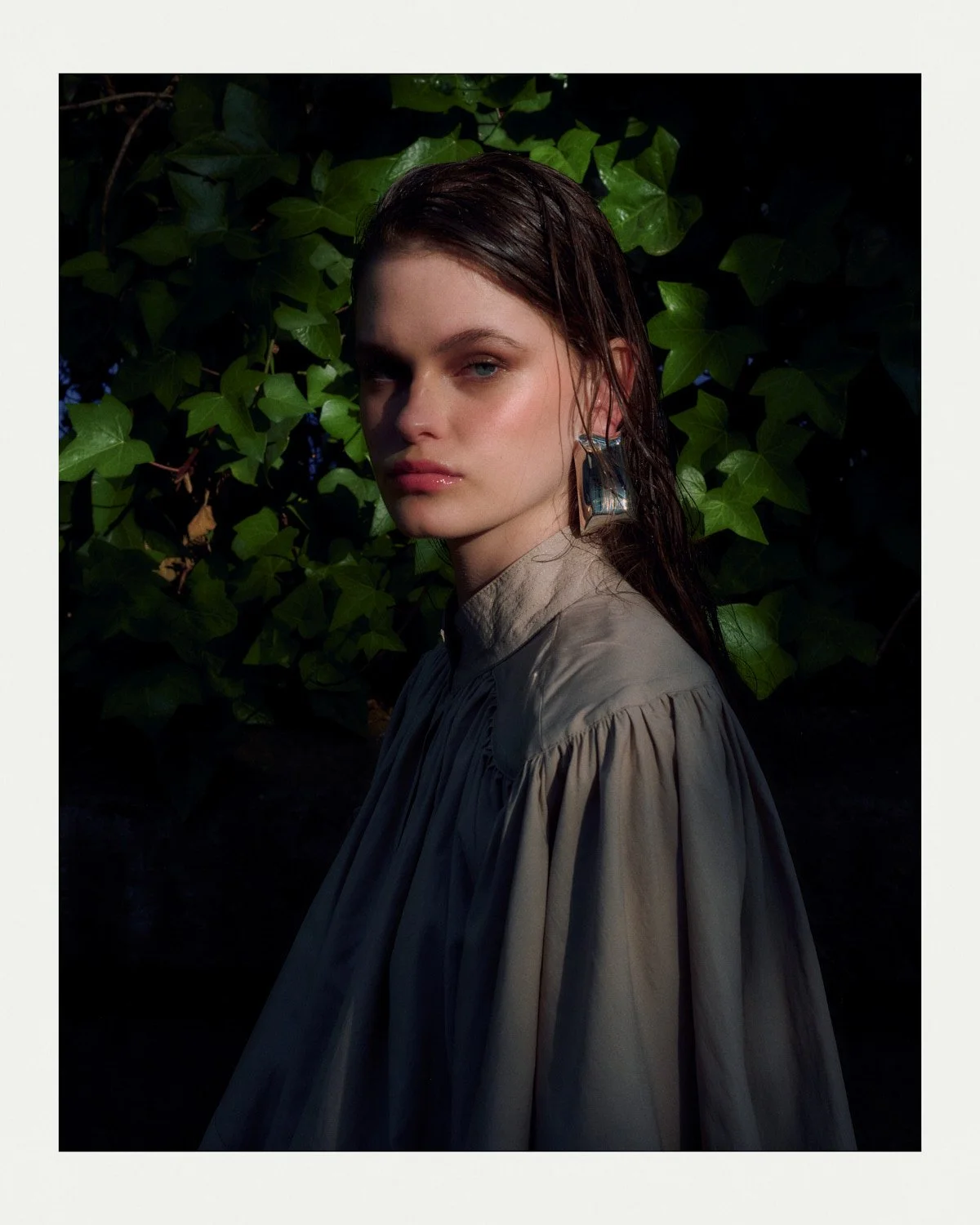 Portrait of a young woman with wet hair, large earrings, and a beige dress, standing in front of green leafy plants with sunlight illuminating her face.