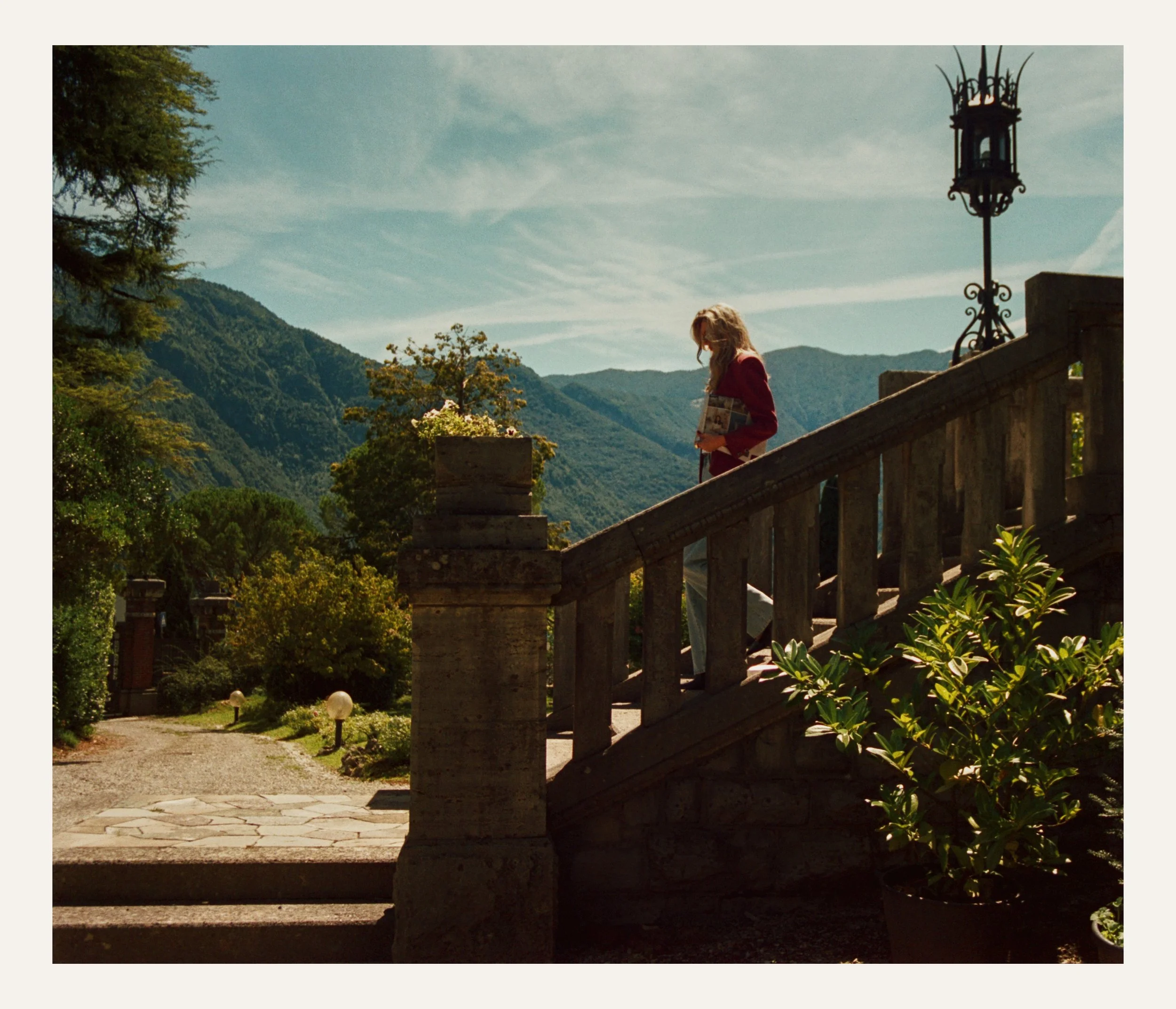 A woman with blonde hair, wearing a red jacket, standing on a stone staircase outdoors, holding a pouch, overlooking a landscape of green mountains, trees, and a partly cloudy sky.