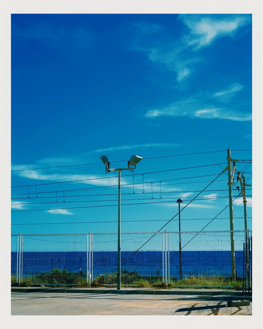 Fencing, electrical wires, and light poles along a coastal road with a view of the ocean and blue sky with scattered clouds.