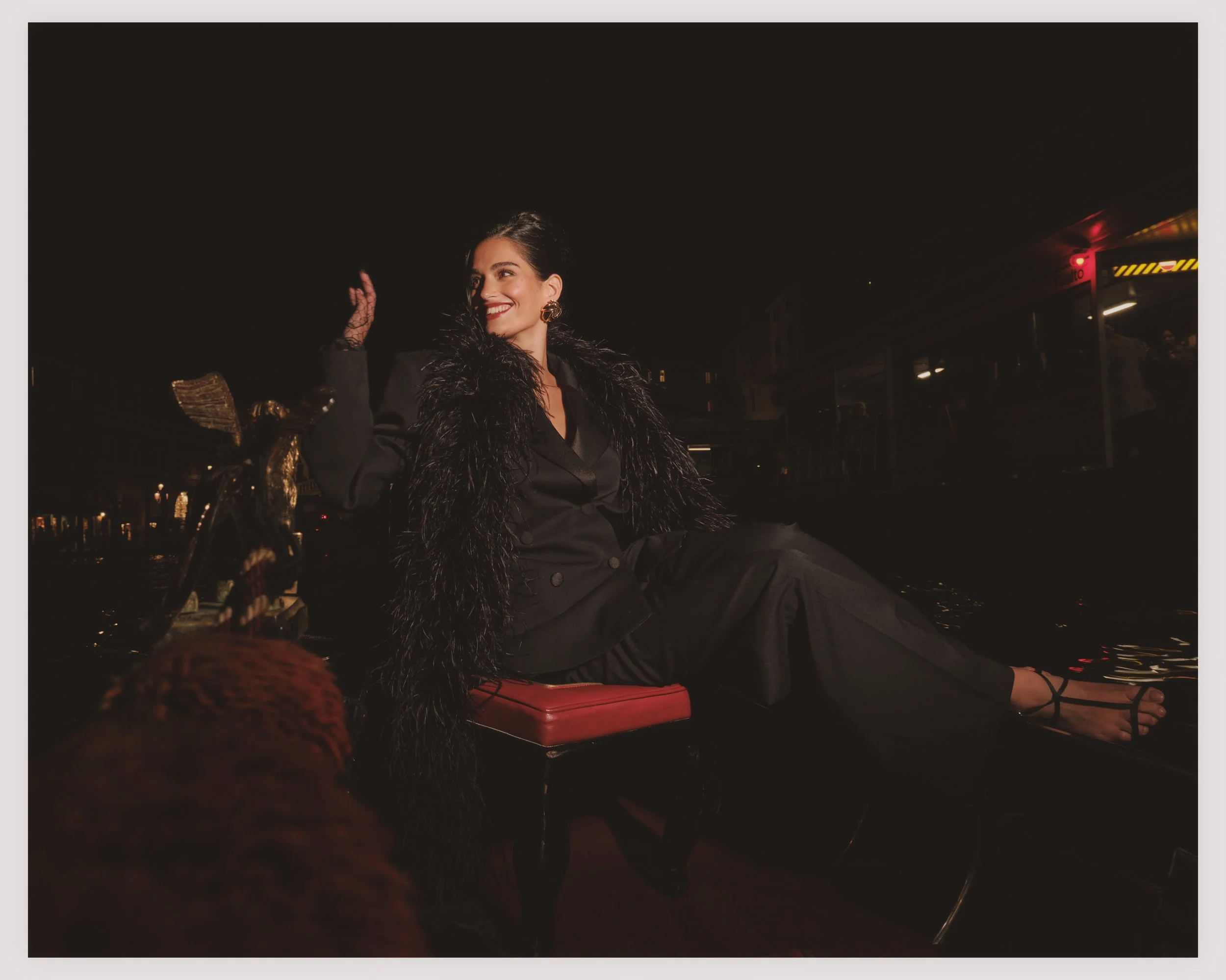 A woman in black clothing with a feathered jacket, sitting on a boat at night, smiling and waving. The background shows city lights and water reflections.