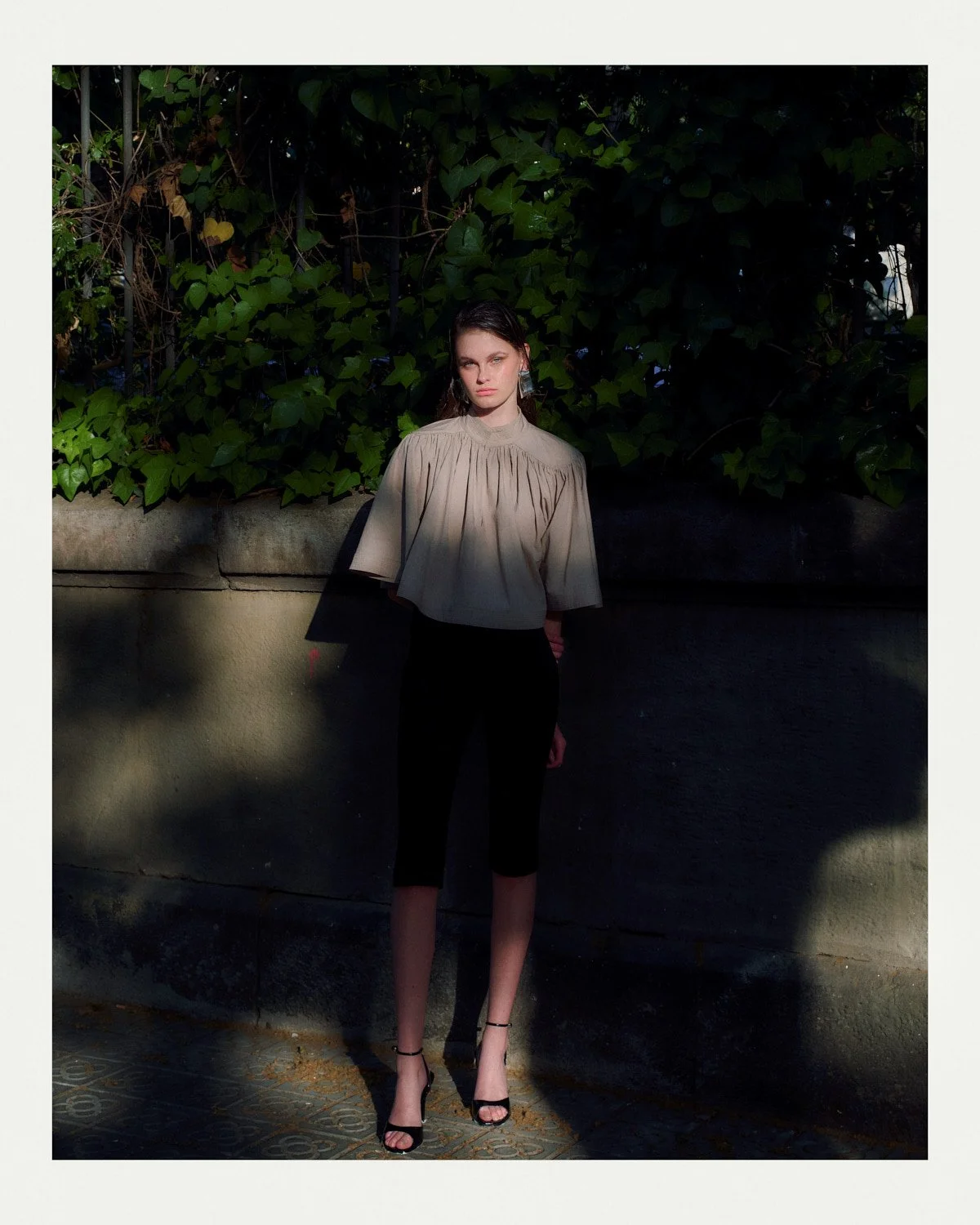A woman standing outdoors against a wall and green foliage, wearing a beige top, black pants, and high heels in sunlight.