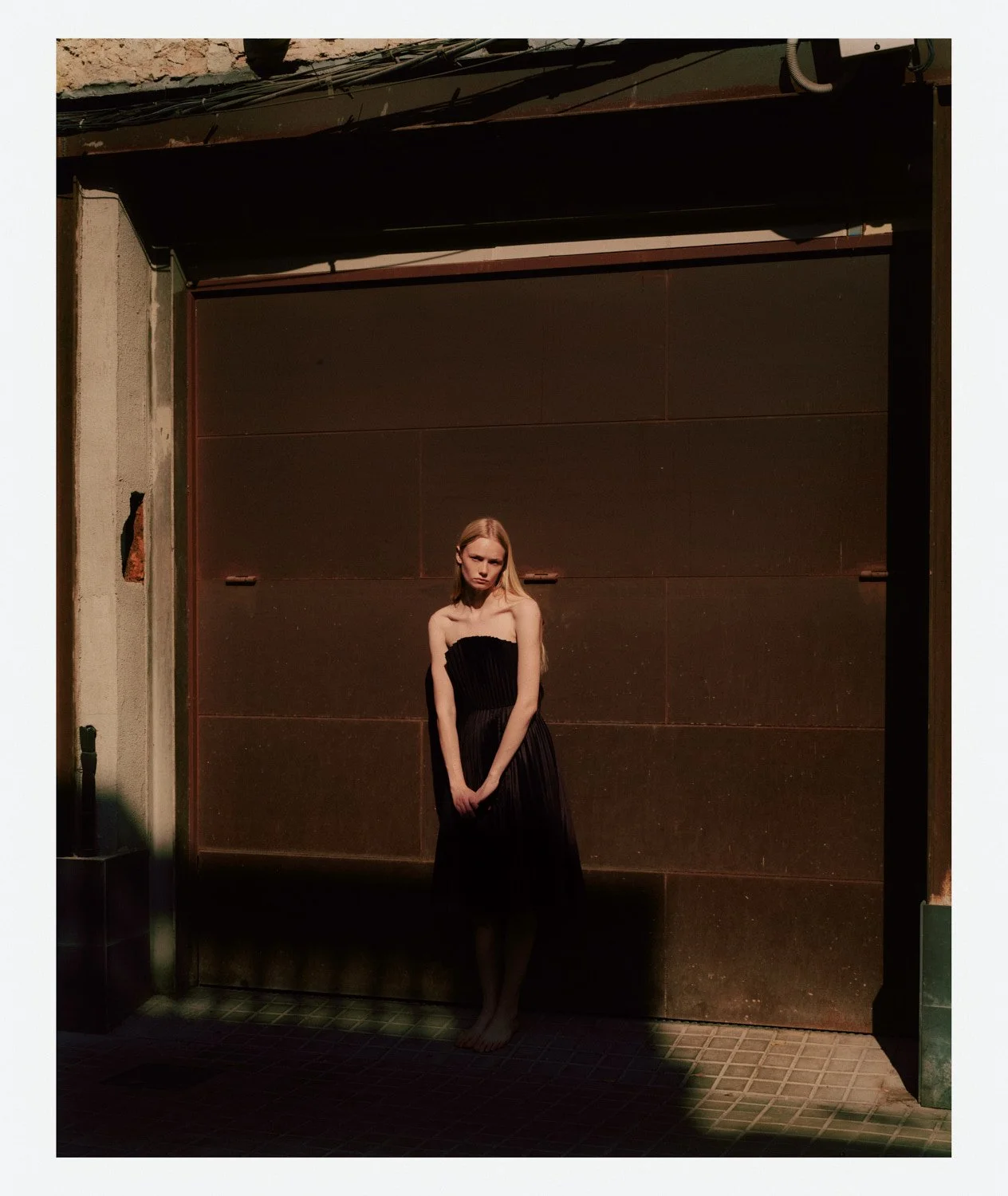 A young woman in a black strapless dress standing barefoot in front of a brown wall with shadows cast on the ground.