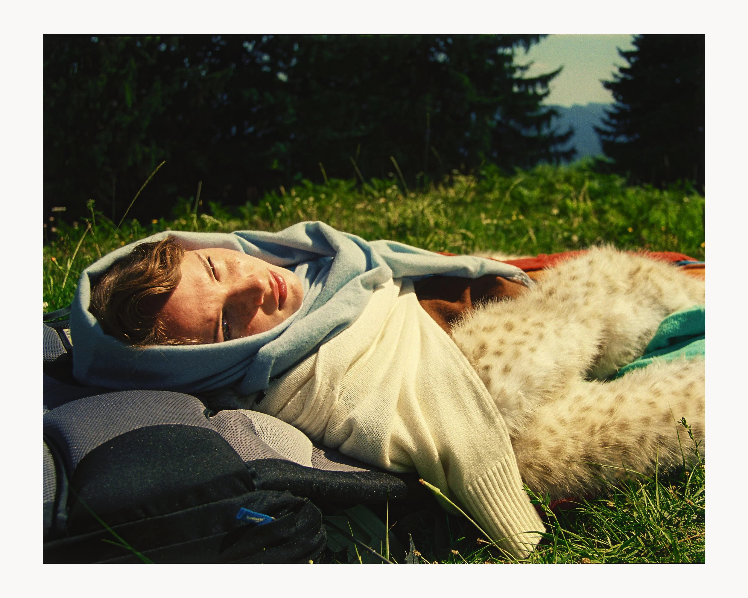 A woman lying on grass with a blanket and gloves, resting outdoors in a forested area.