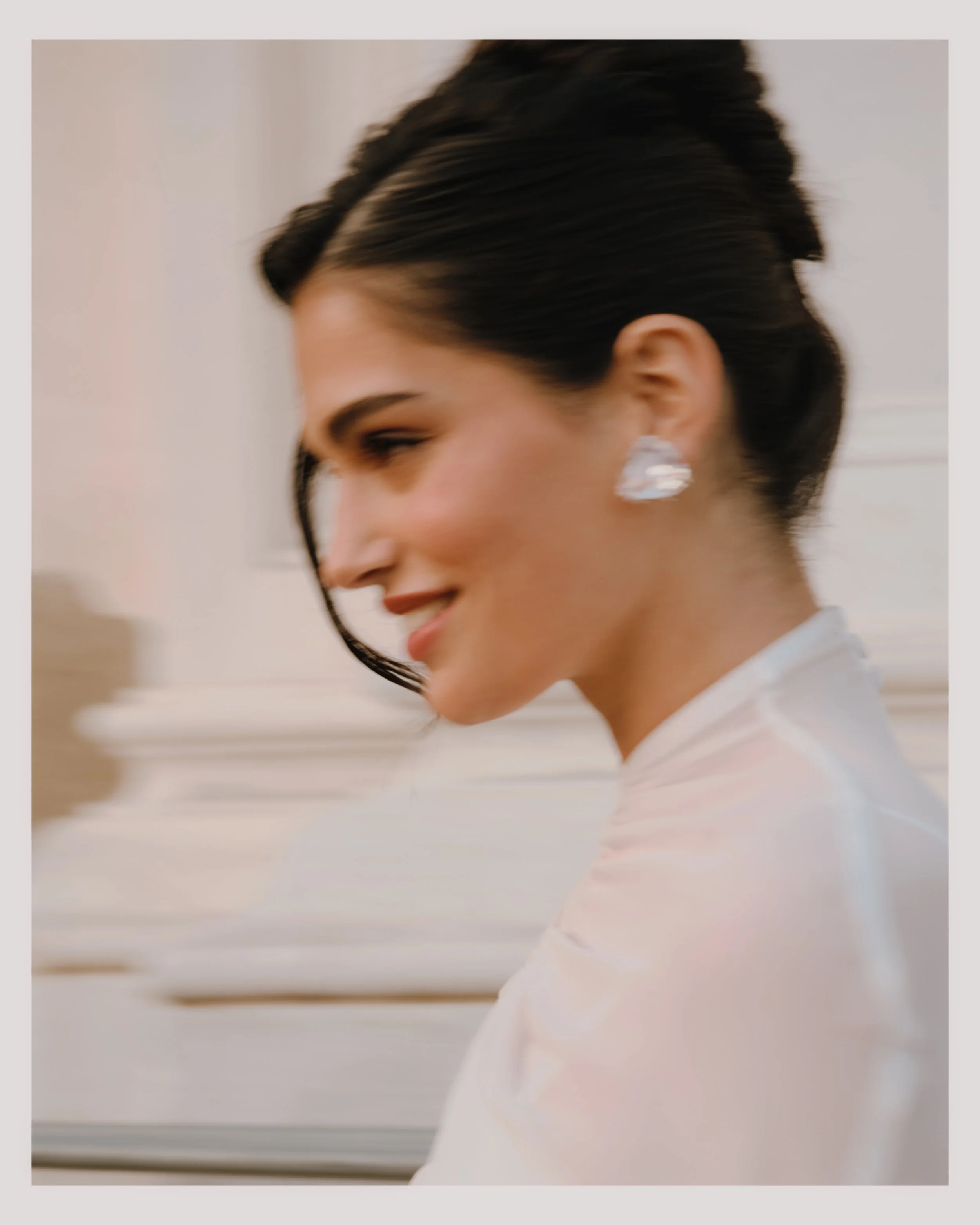 Close-up of a woman with short dark hair, earrings, and a white top, smiling softly and looking to the side, with a blurred white background.