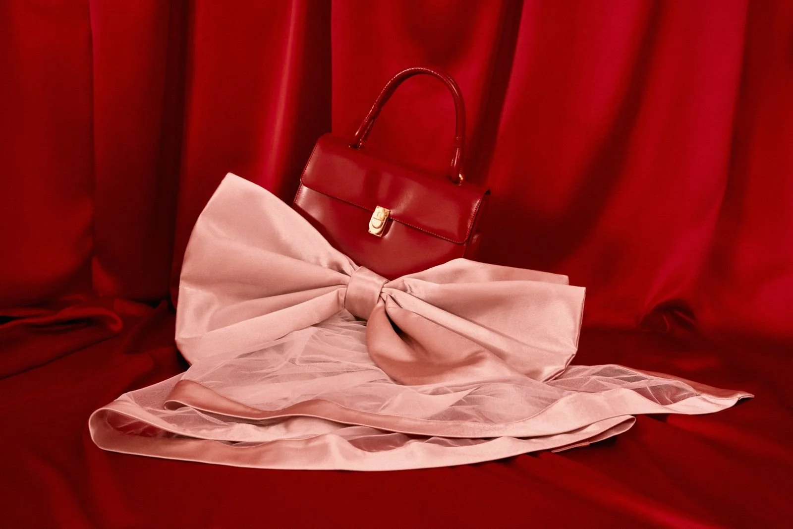 A red handbag with a gold clasp, placed on red curtains with a pink silk scarf tied in a large bow, all arranged on a red fabric surface.