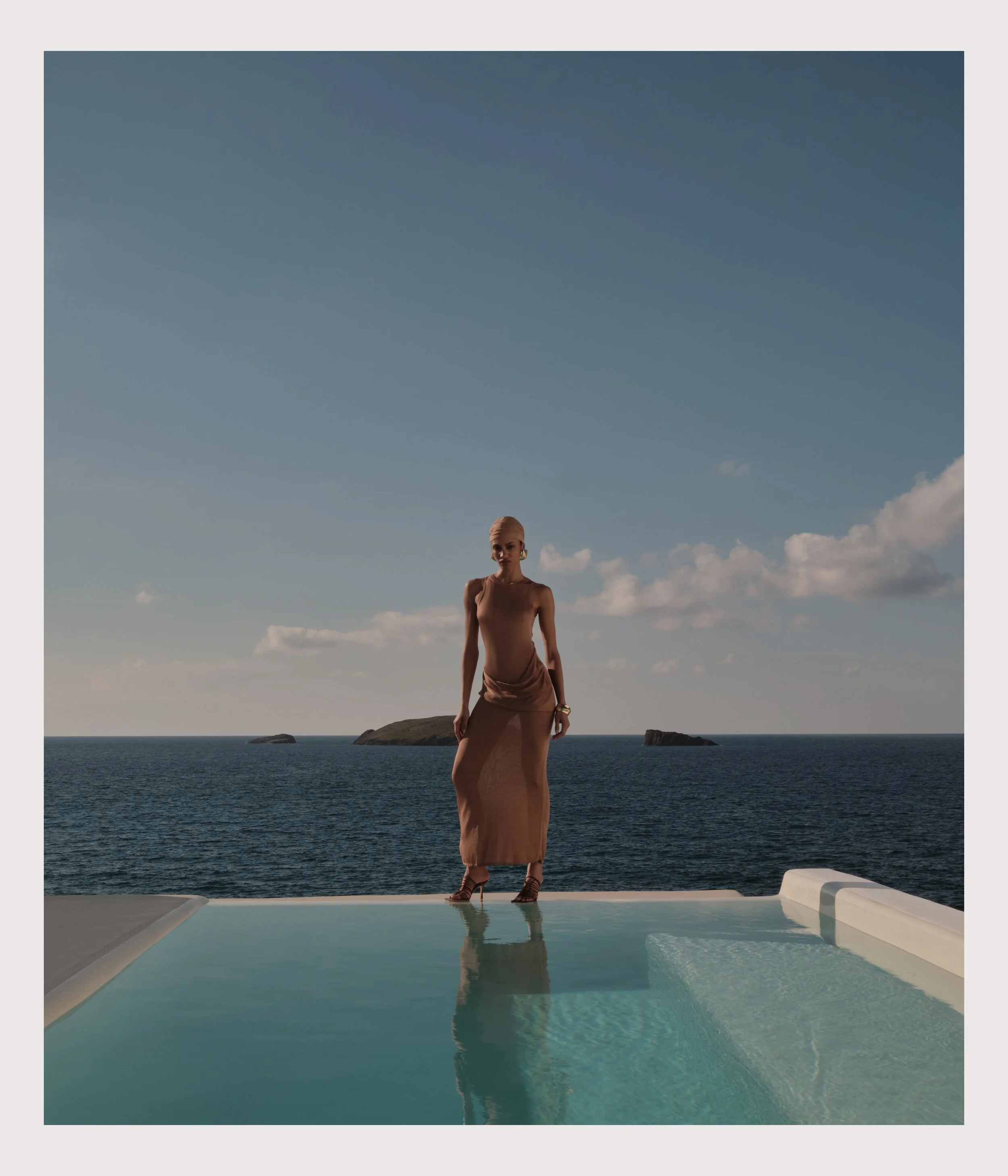 A woman in a brown dress standing on the edge of a swimming pool with the ocean and distant islands in the background under a sky with few clouds.