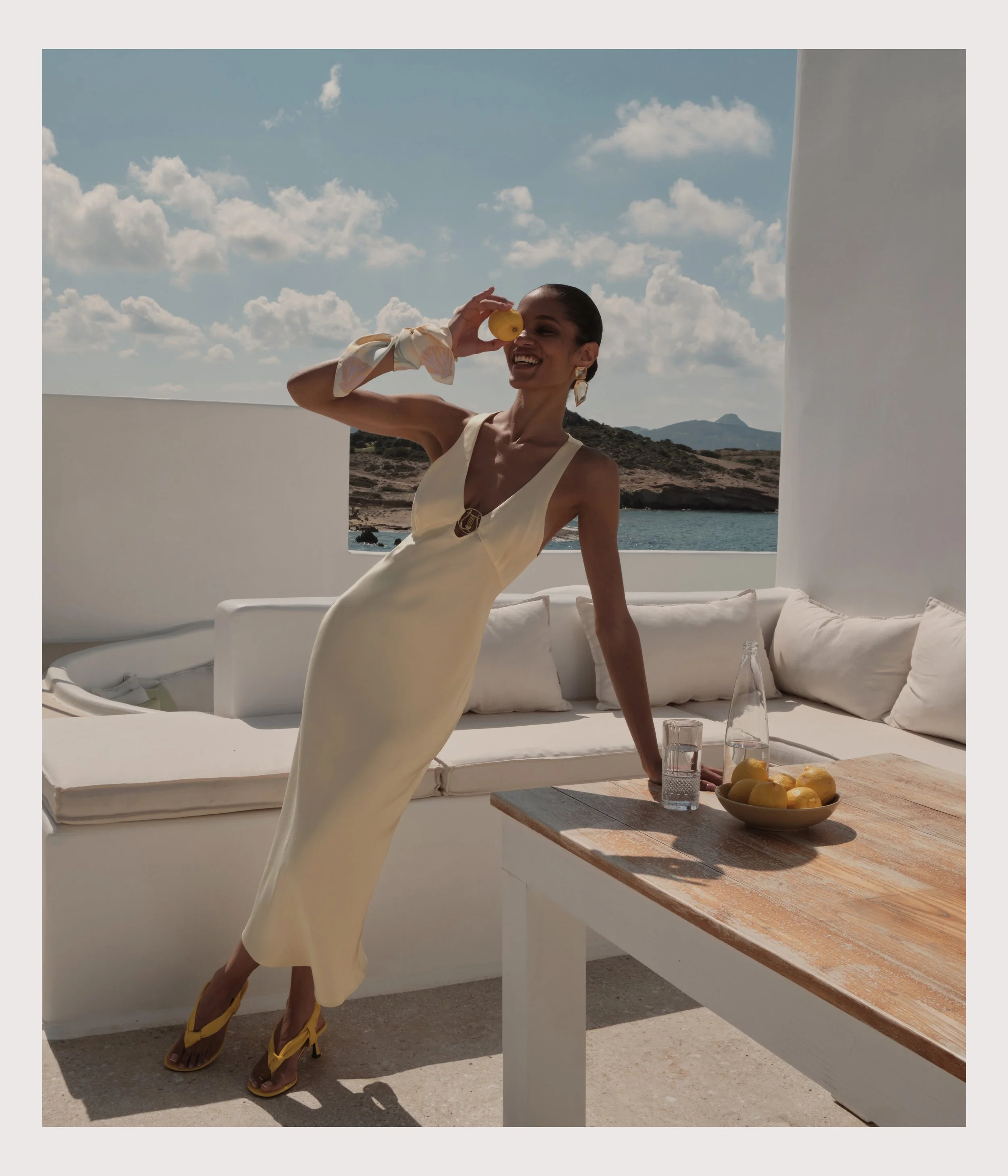 A woman in a yellow dress and matching yellow heeled sandals standing on an outdoor patio, holding a lemon near her eye, with a scenic coastal background and white patio furniture and table with bowls of lemons.