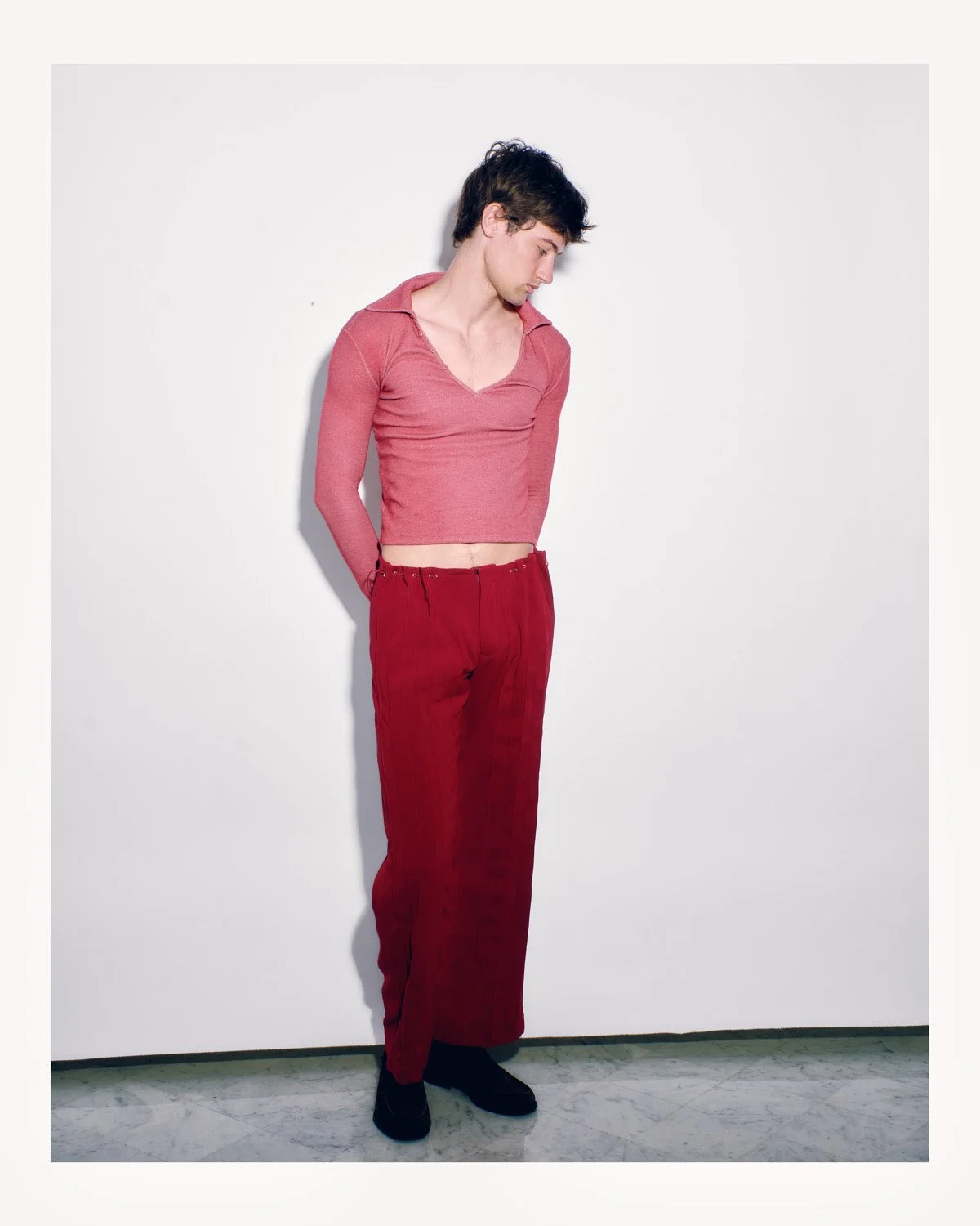 A young man with dark hair, wearing a pink cropped long-sleeve shirt and red pants, stands against a plain white background, looking downward with his hands behind his back.