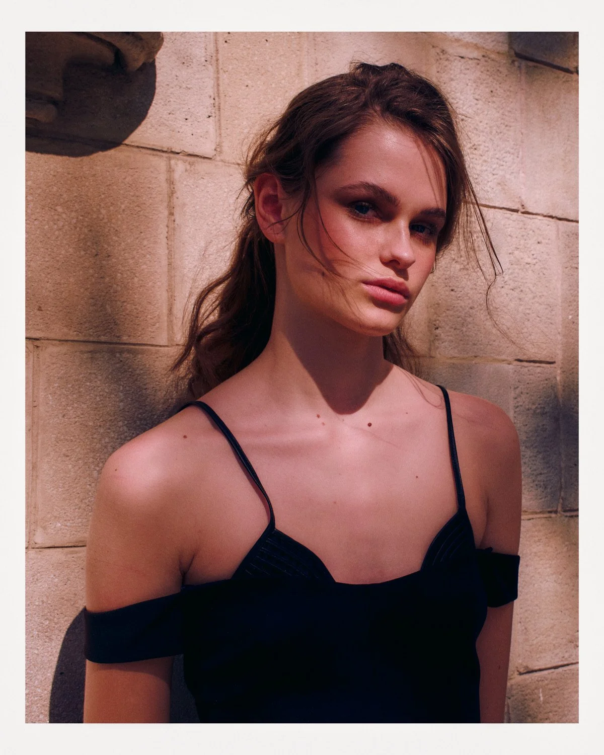 A young woman with light skin, brown hair, and blue eyes, wearing a black spaghetti strap top, standing against a beige brick wall with shadow patterns.