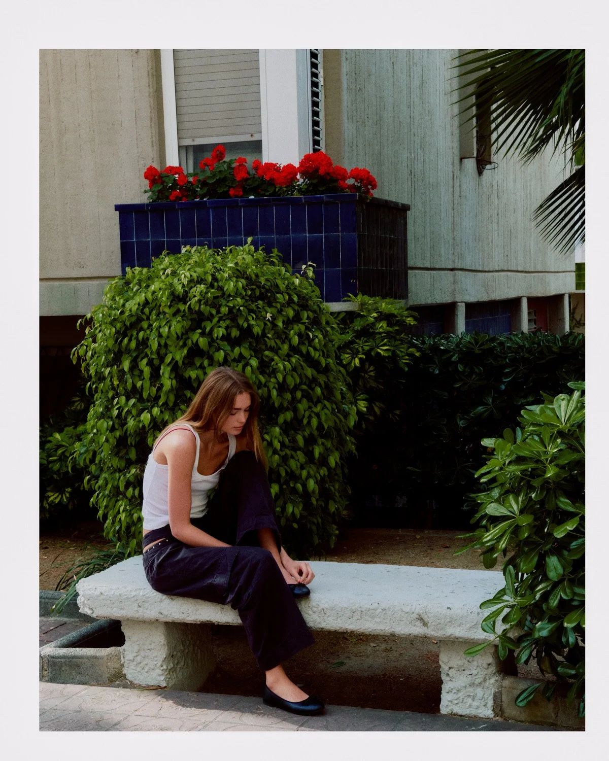 A young woman with long hair, wearing a white tank top and black pants, sitting on a white concrete bench outdoors surrounded by green bushes and plants. A building with a blue planter filled with red flowers is in the background.