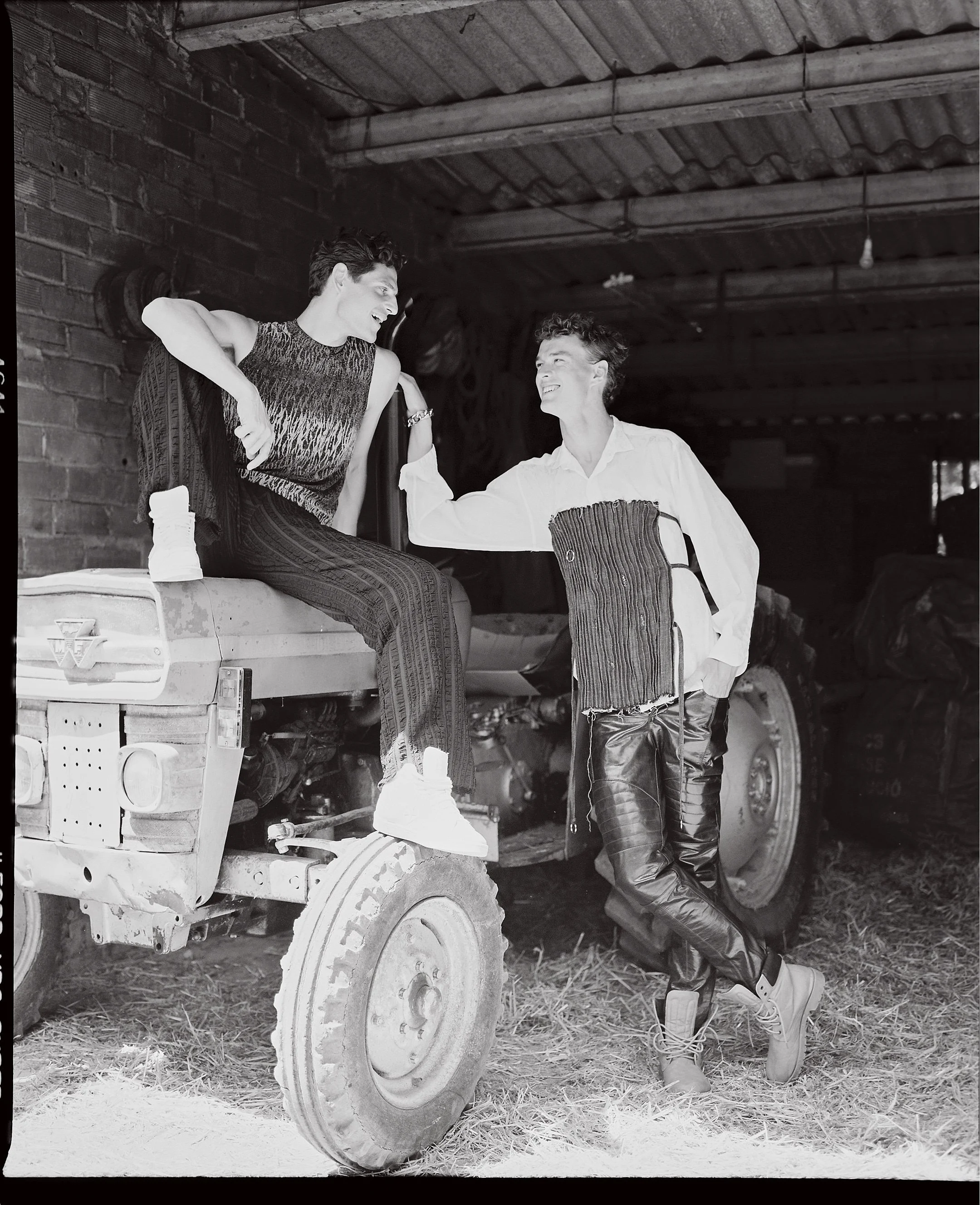 Two young men having a friendly conversation in a rustic barn. One is sitting on a tractor, and the other is standing next to him, leaning on the tractor. The scene is casual and cheerful.