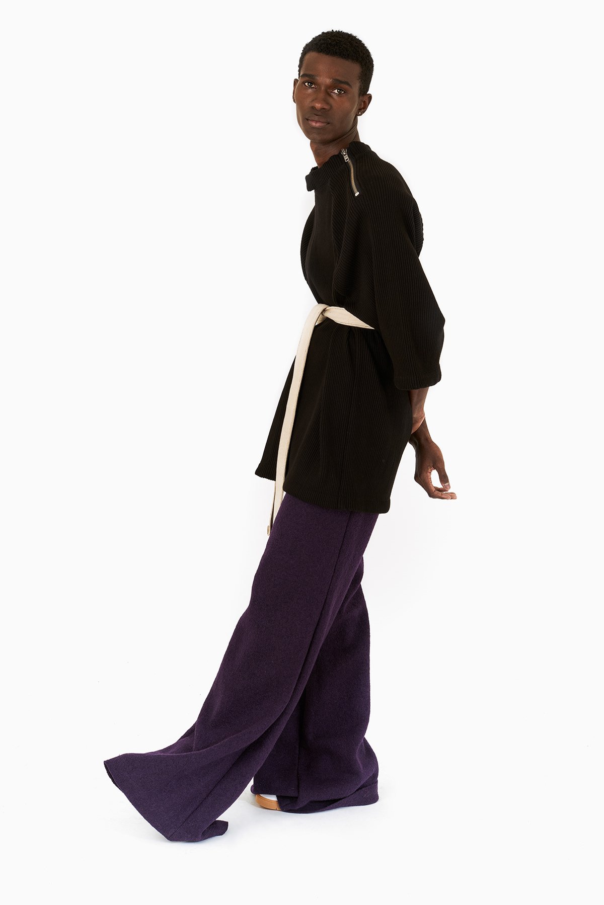 A young man with dark skin and short curly hair standing against a white background, wearing a black oversized jacket with zipper details, purple wide-leg pants, and a white belt, looking directly at the camera.