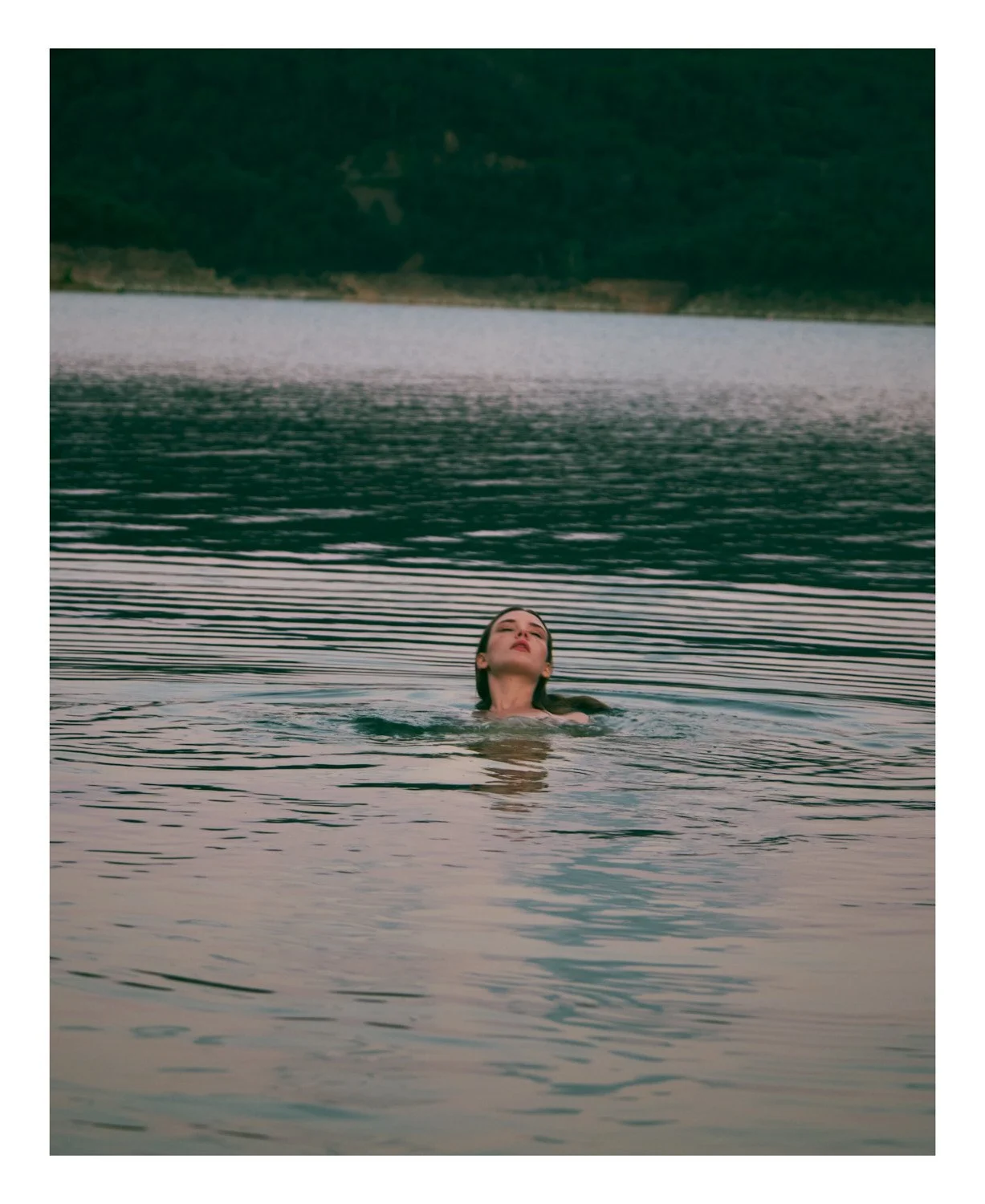 A woman with dark hair swimming in a lake, with mountains in the background and overcast sky.