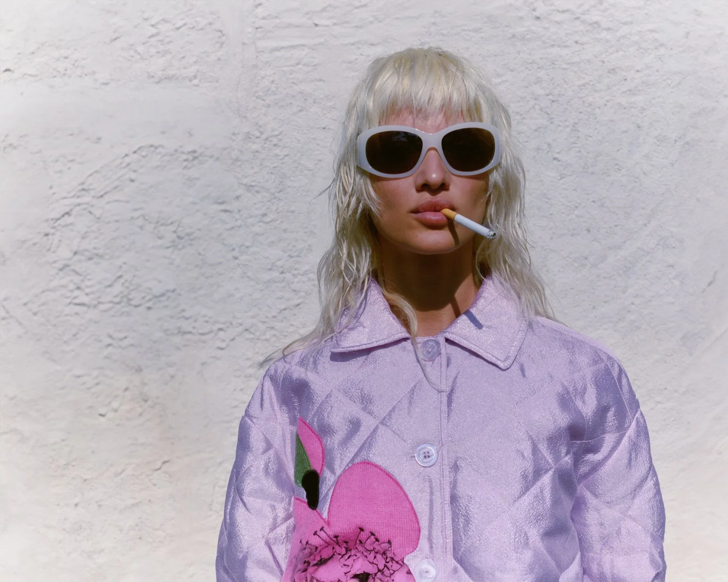 A woman with long, wavy blonde hair wearing large white sunglasses, a lavender jacket with a pink heart and green leaf embroidery, and smoking a cigarette against a textured white wall.