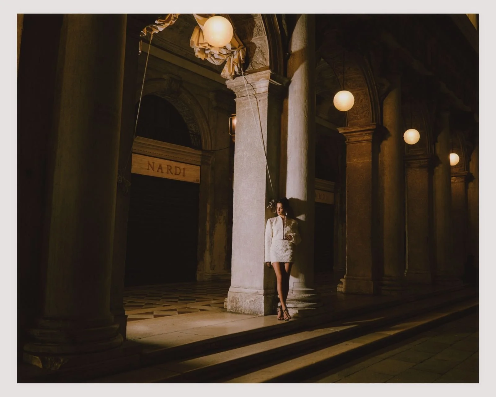 A woman in a white dress and high heels standing next to a large column in a dimly lit, ornate architectural setting with arches and hanging globe lights.
